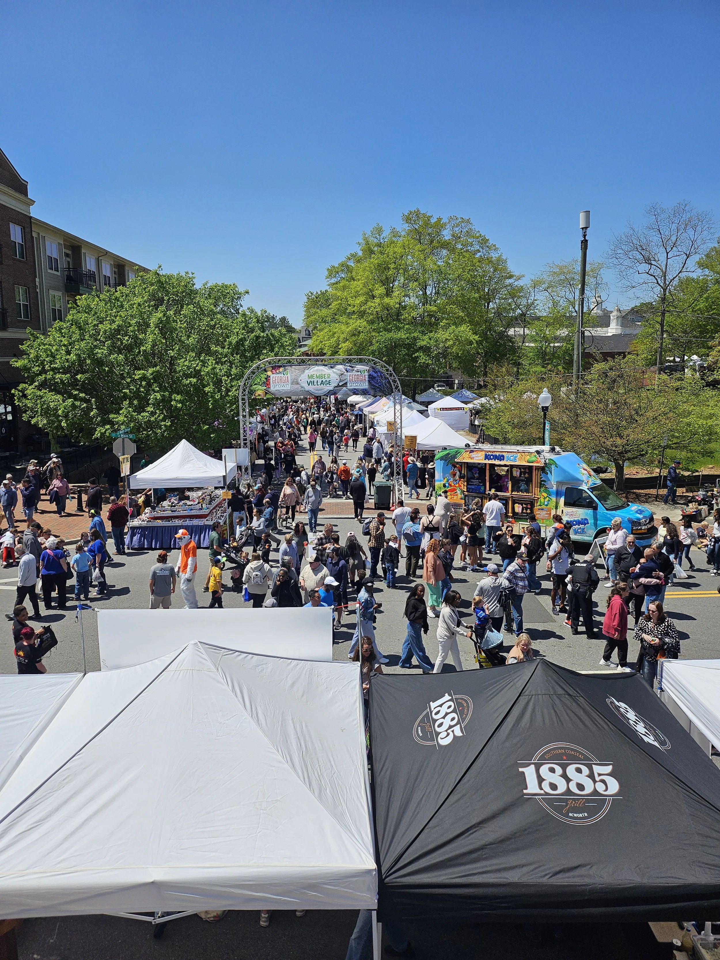 A street fair with numerous tents and booths, filled with people walking and browsing under a clear blue sky, surrounded by green trees and nearby buildings.