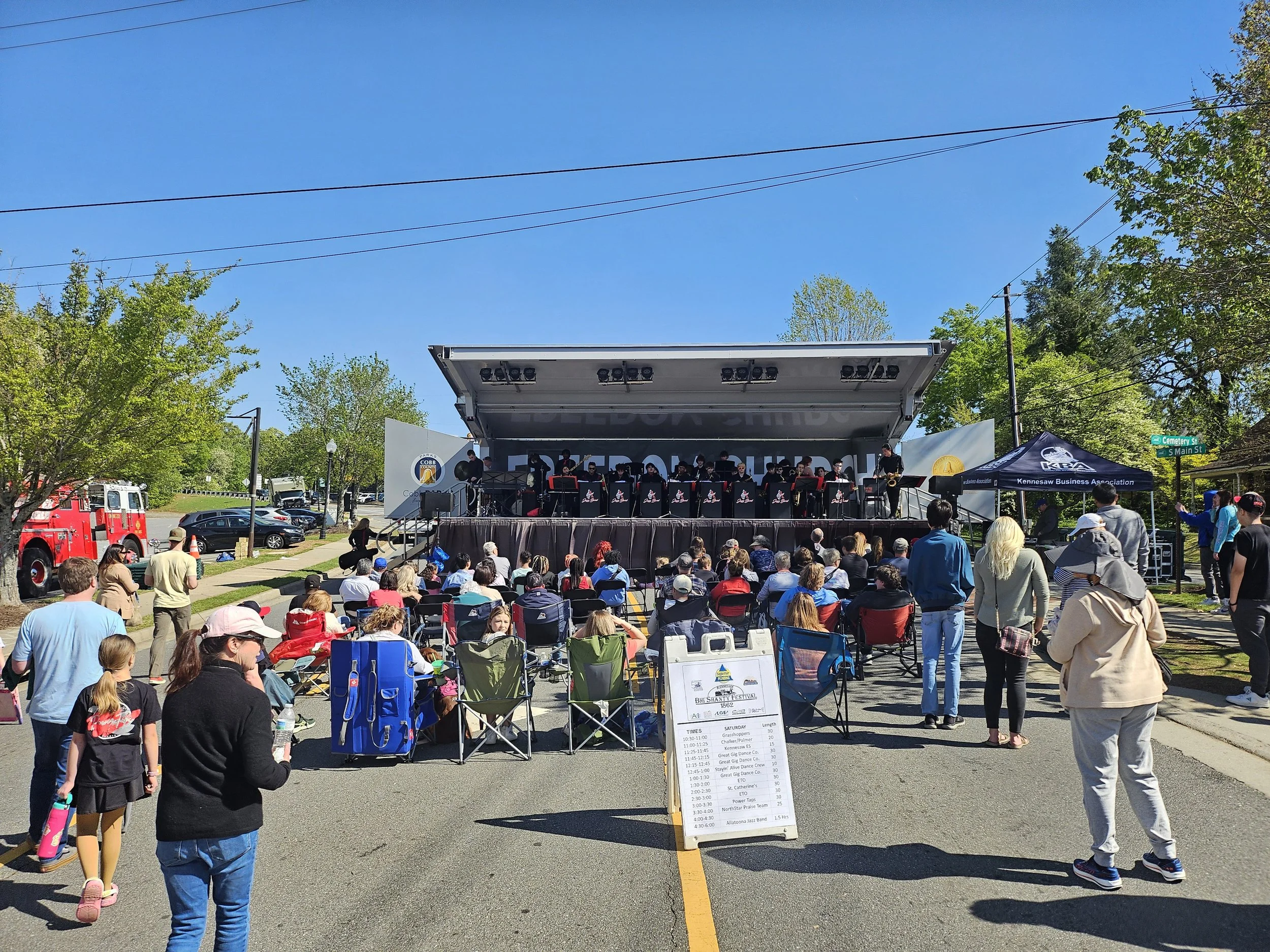 Outdoor community event with a live music band on stage, audience seated in chairs, and people walking around on a sunny day.