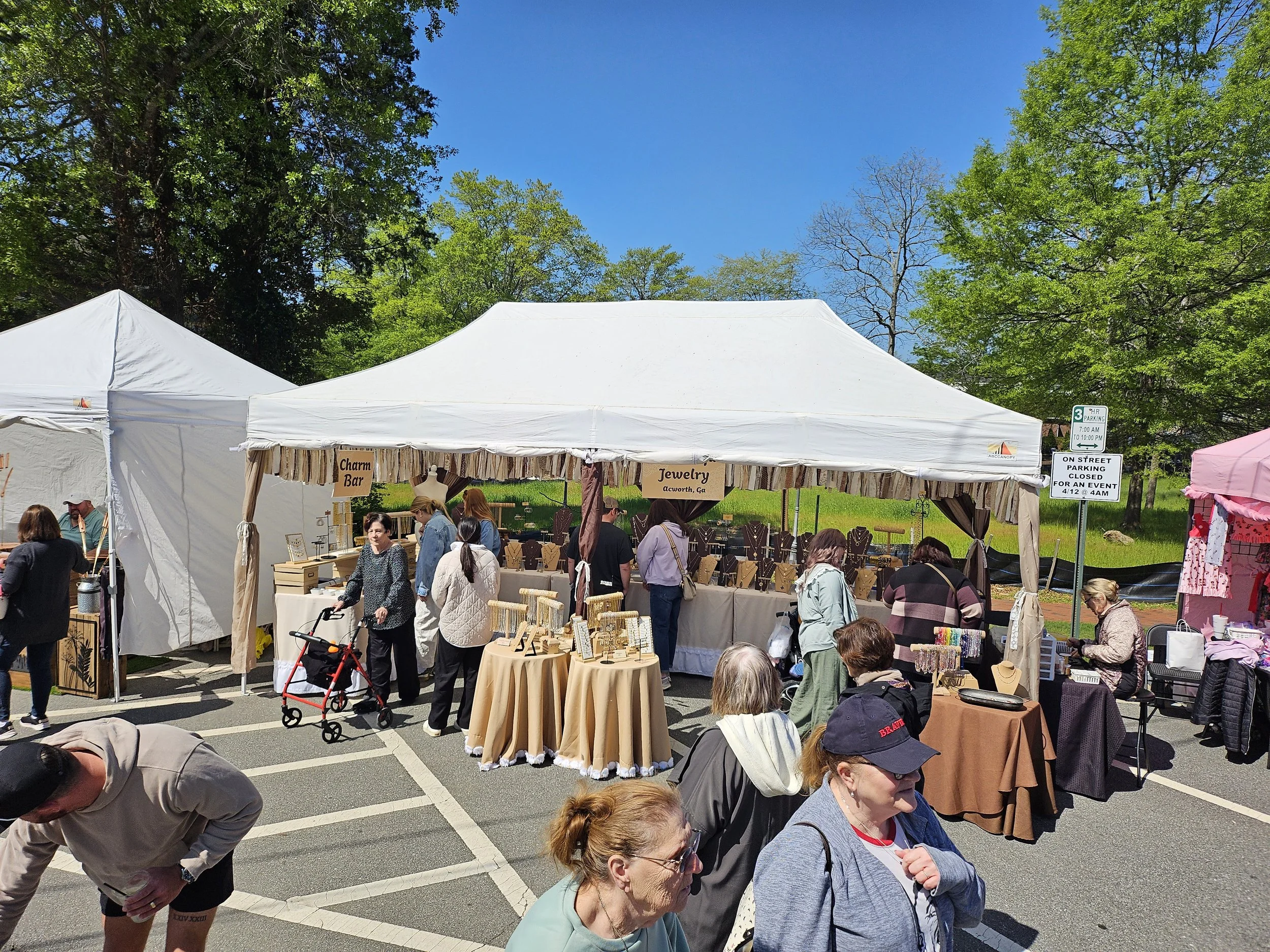 Outdoor market scene with vendors under white tents selling jewelry and charm items, and shoppers browsing and walking in parking lot with trees in the background.