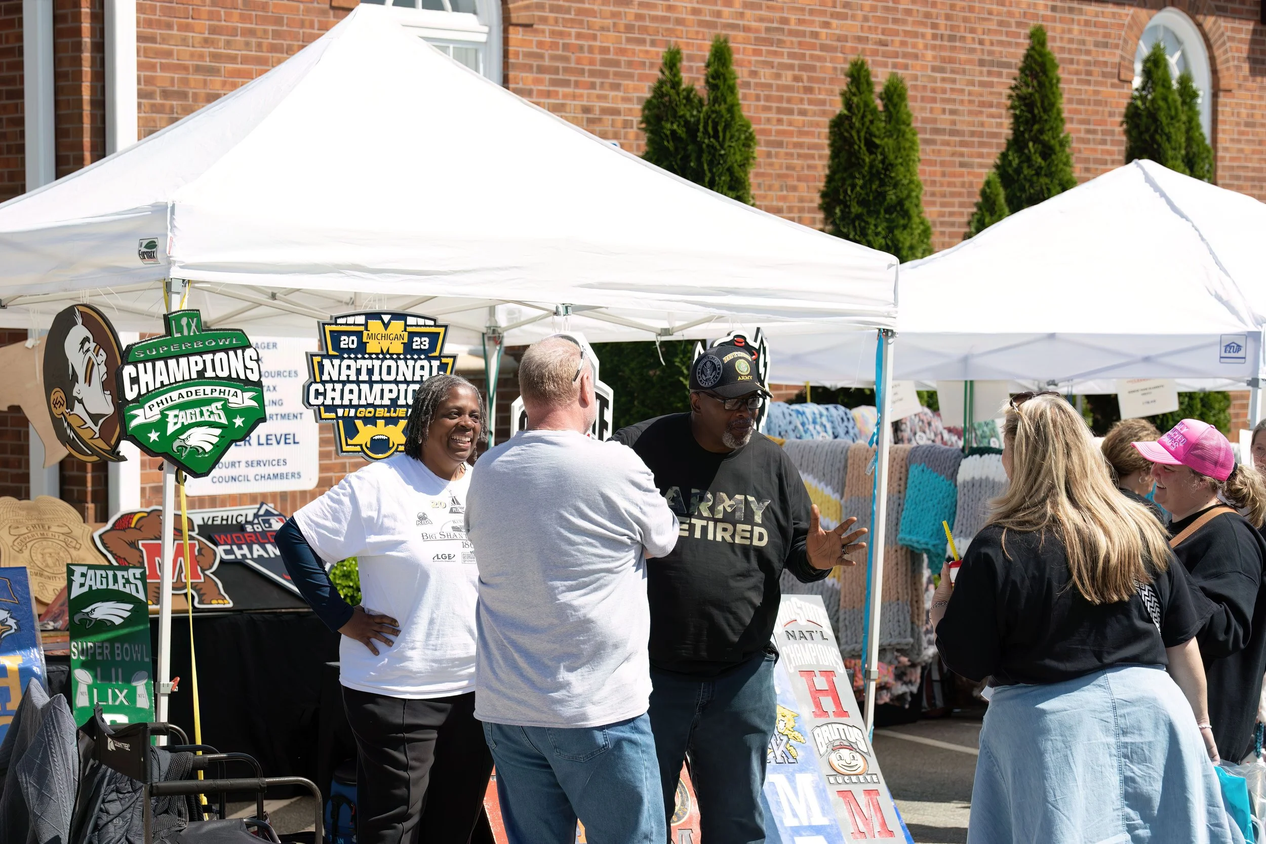 People talking at an outdoor booth with sports memorabilia, including signs for Eagles Super Bowl Champions and Michigan National Champions, with a brick building and trees in the background.