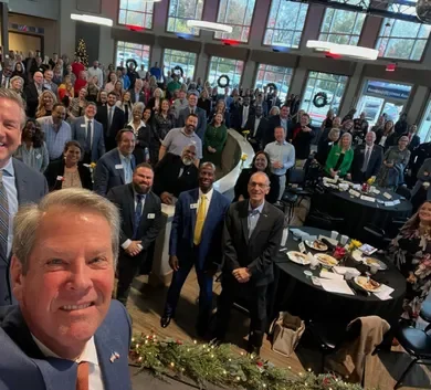 A group of people in a large indoor event space, many dressed in formal or business attire, posing for a group photo during a celebration or conference. There are tables set with food and decorations, and large windows letting in natural light.