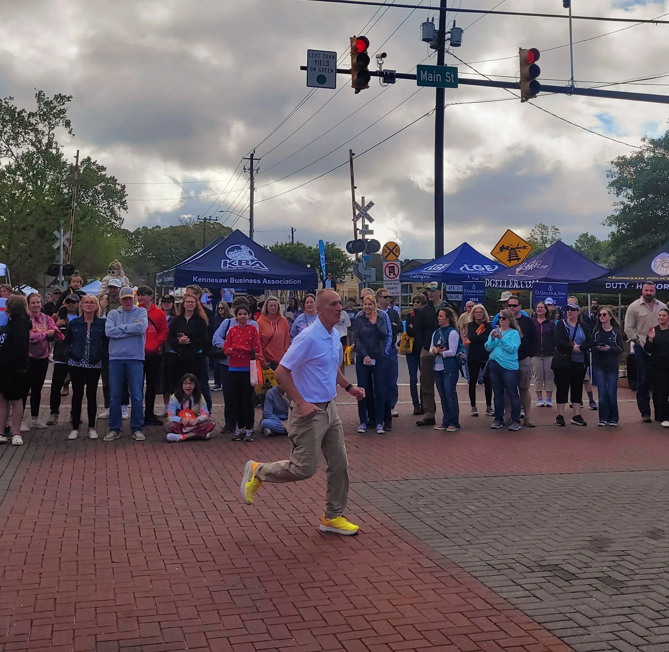 A man jogging across a brick street while a crowd gathers behind him for an outdoor event with tents and banners. Overhead traffic lights and street signs are visible.