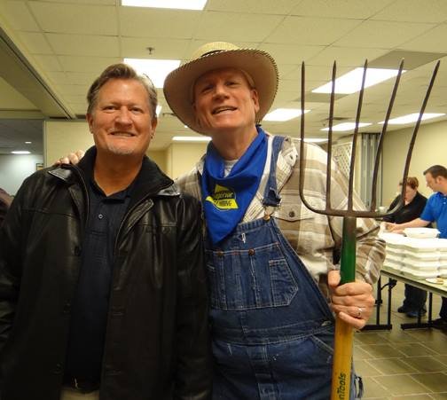 Two men smiling indoors; one wearing a black leather jacket, the other dressed as a farmer with a straw hat, overalls, and holding a pitchfork.
