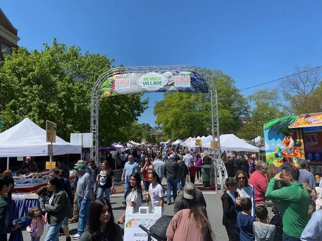 People walking under a metal archway at an outdoor fair or festival with tents and trees around.