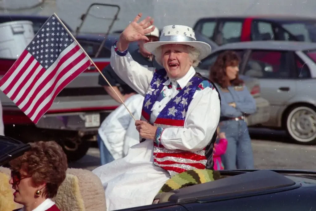 An elderly woman dressed in patriotic clothing, waving an American flag, riding in a convertible car during a parade, with onlookers in the background.