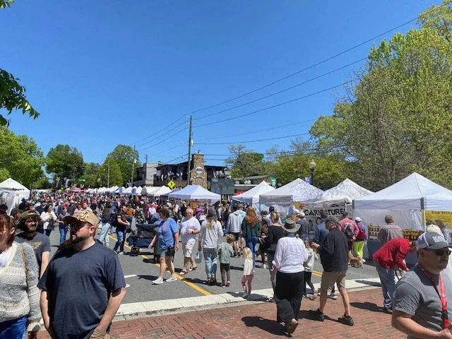 A busy outdoor market or fair with white tents and numerous people walking and browsing on a sunny day with clear blue skies.