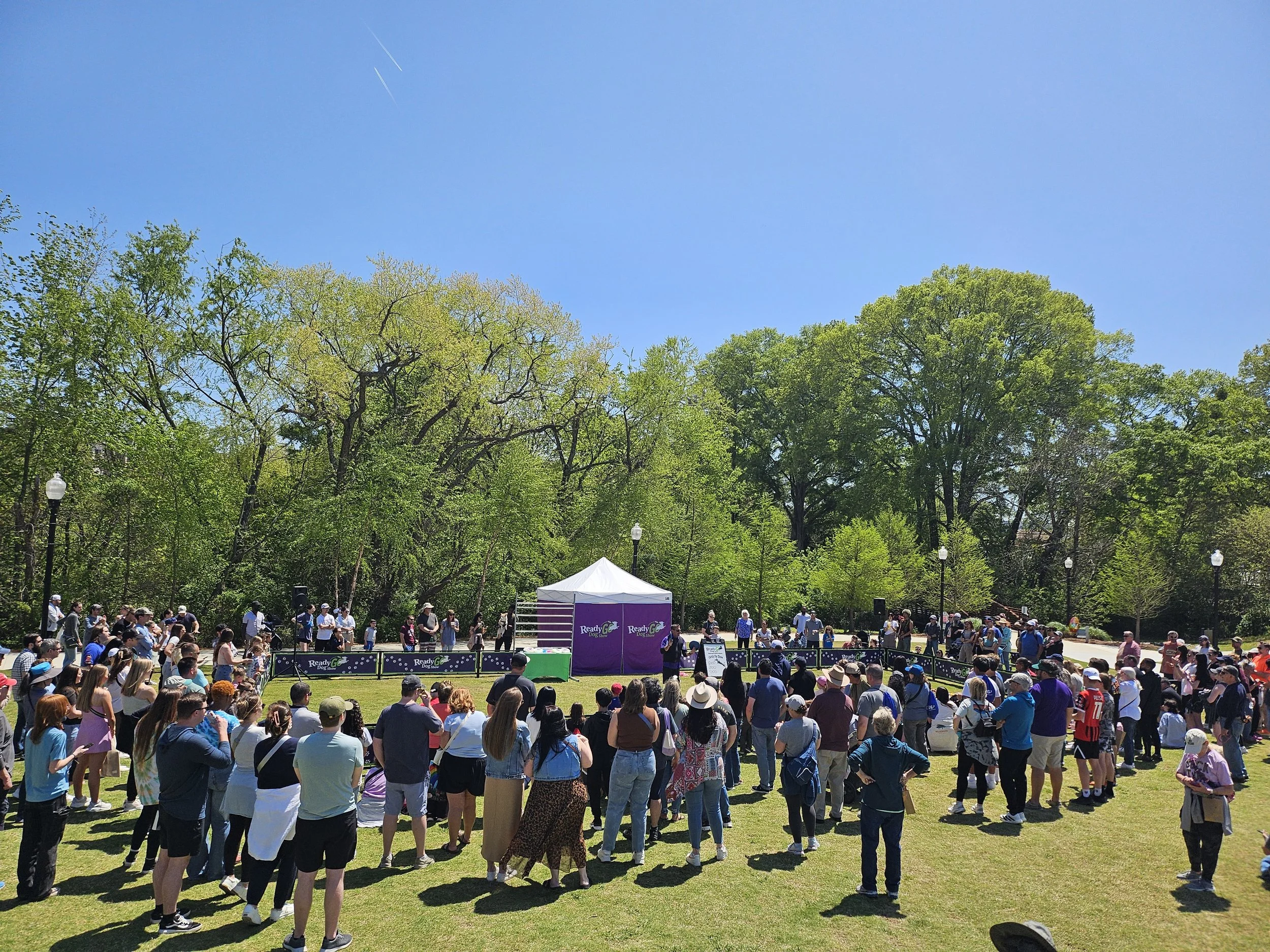Crowd gathered outdoors at a public event under a clear blue sky, with a white tent and a stage in the background surrounded by green trees and lamp posts.