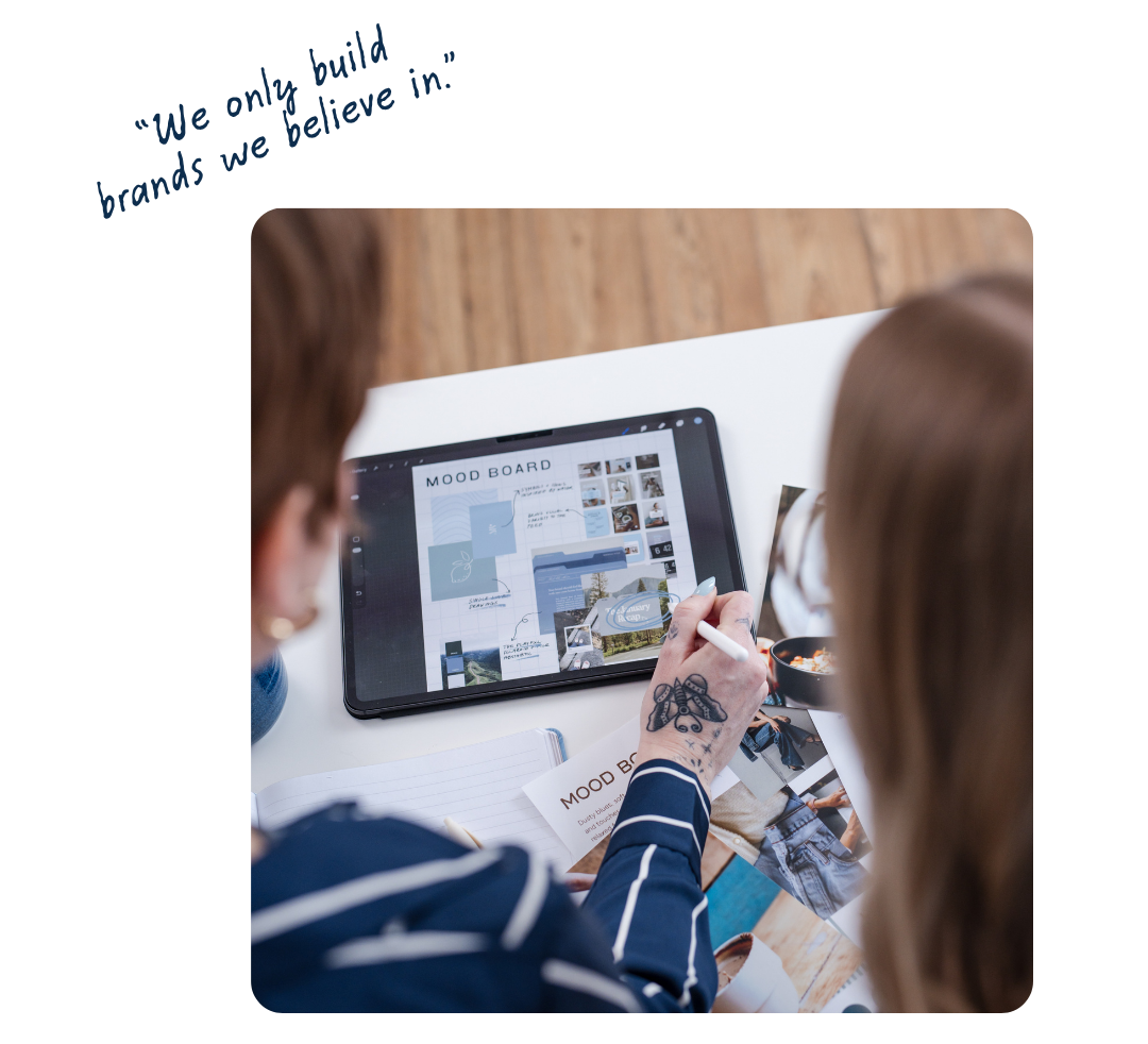 Two people are working together on a mood board using a tablet. One person is pointing at the screen, which displays images and notes related to lifestyle or design planning. The table has printed materials and a notebook.
