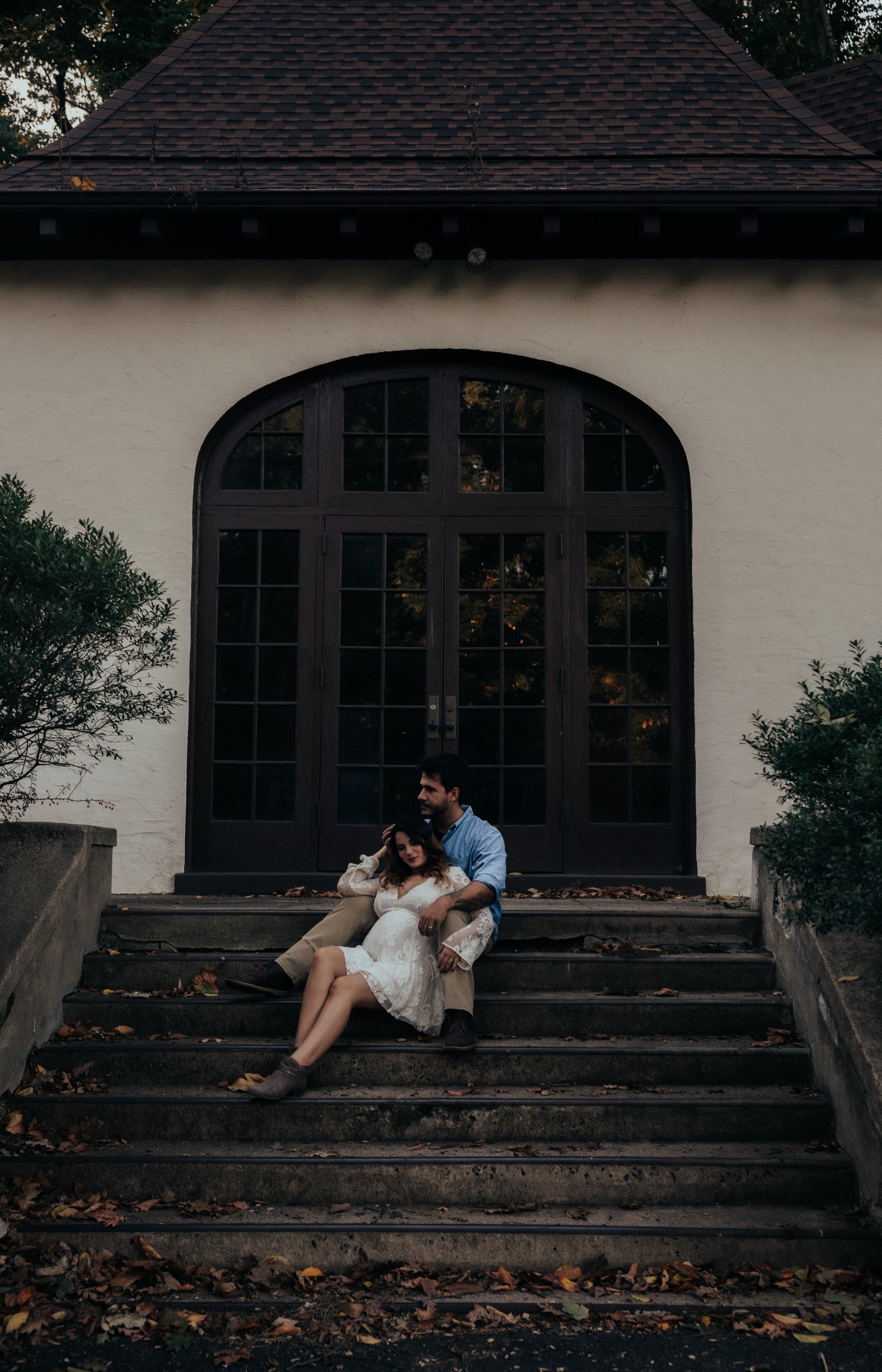 A couple sitting on outdoor stairs in front of a large dark-framed glass door, with trees and fallen leaves around, during evening.