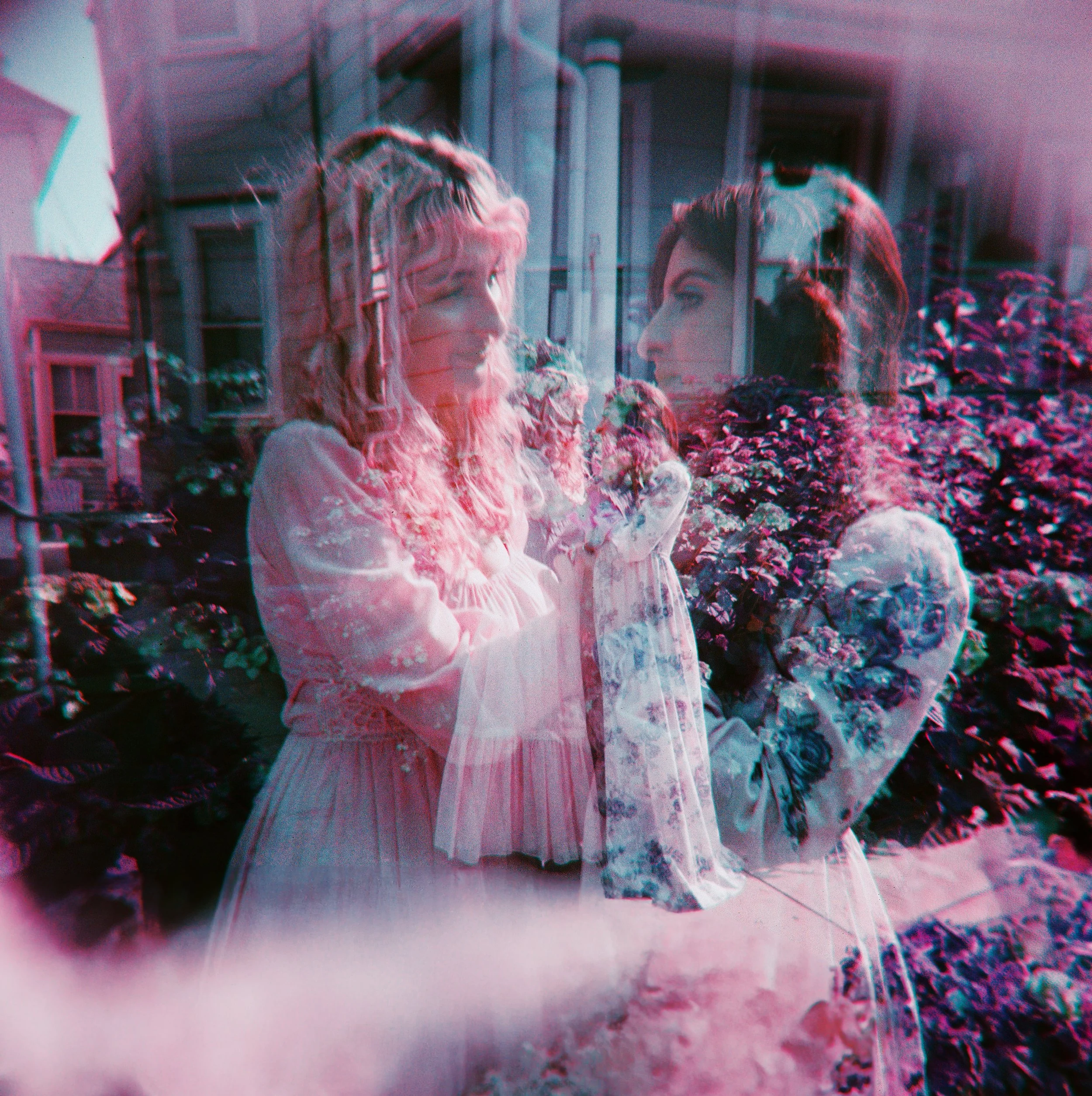 A double exposure photograph of two women with long hair, wearing long dresses, surrounded by plants and flowers in an outdoor garden or porch area.