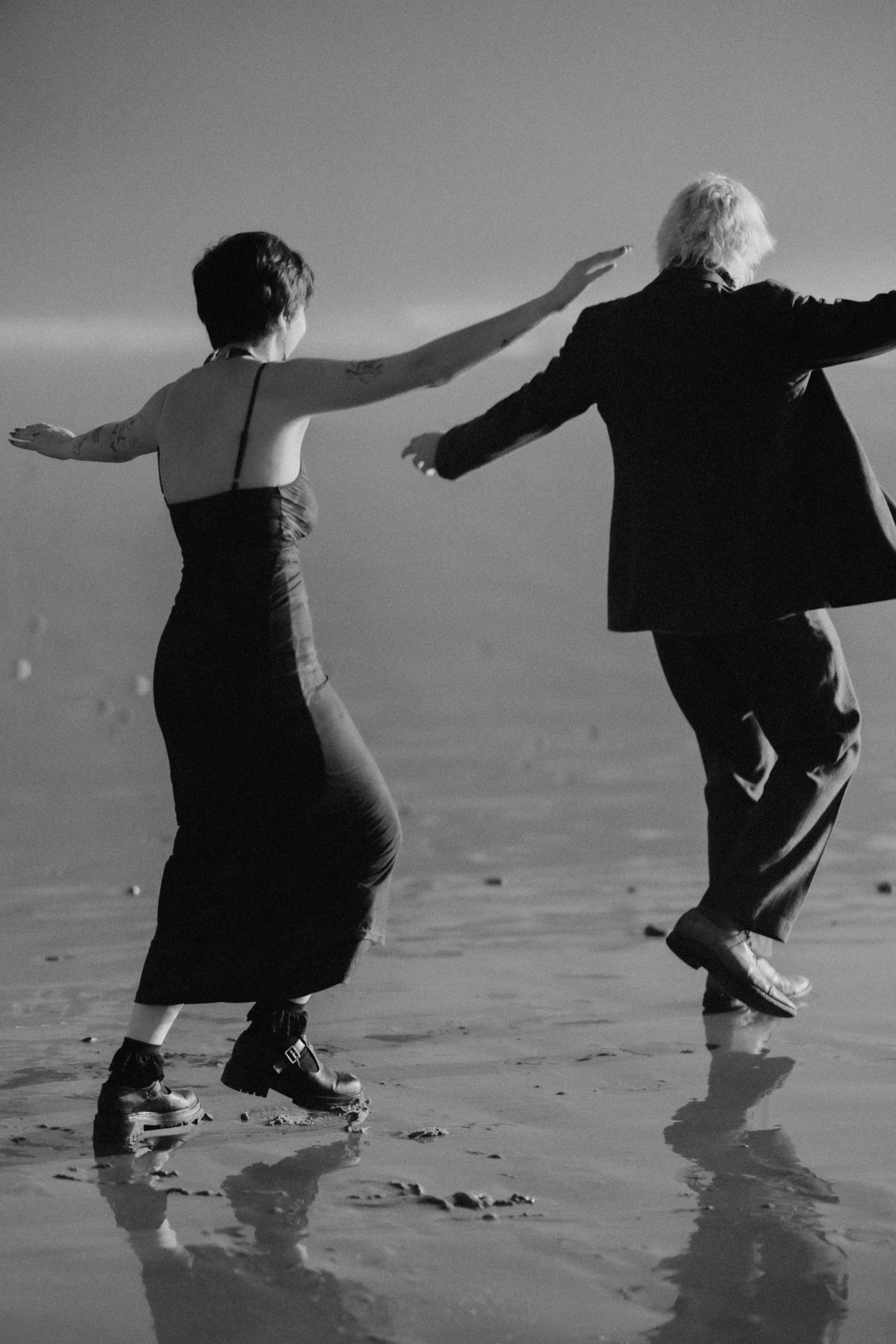 Two people dancing on the beach, with one person wearing a long dress and the other in a suit, captured in black and white.