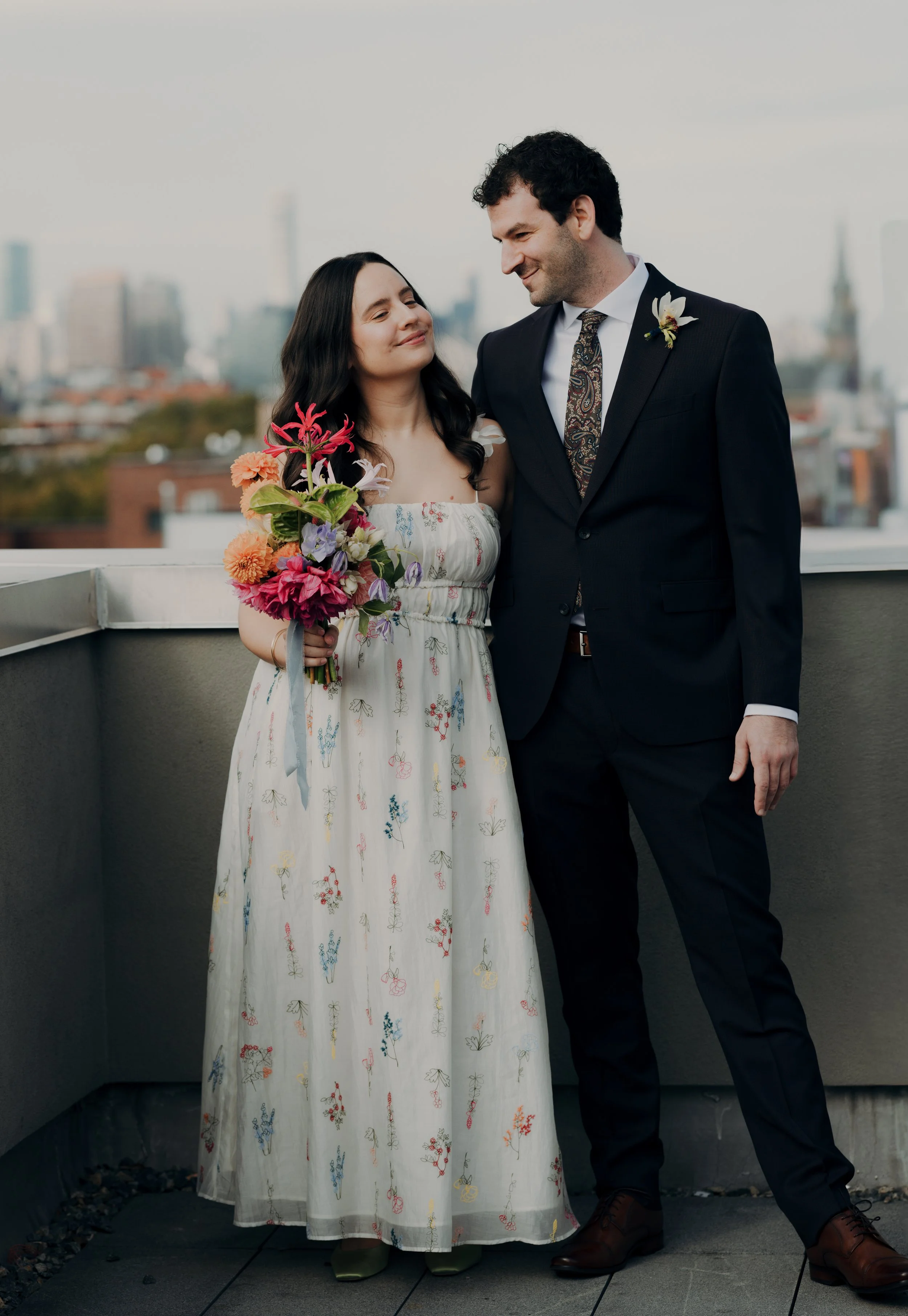 A couple standing on a rooftop, with city buildings in the background. The woman is holding a colorful bouquet and wearing a white floral dress, while the man is dressed in a dark suit with a boutonniere.