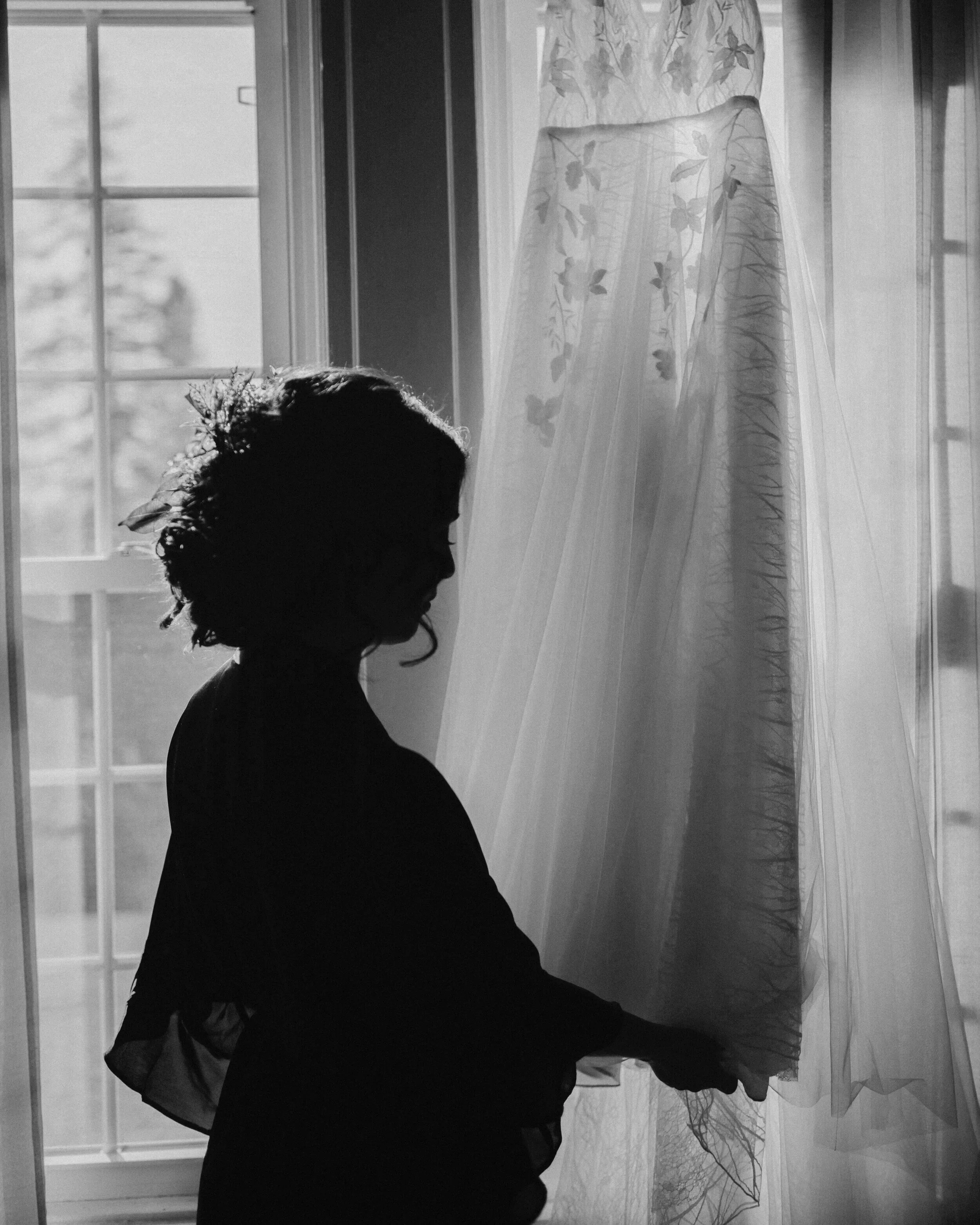 Silhouette of a woman with curly hair, standing in front of a window with sheer curtains, holding the curtain with one hand, backlit by natural light.