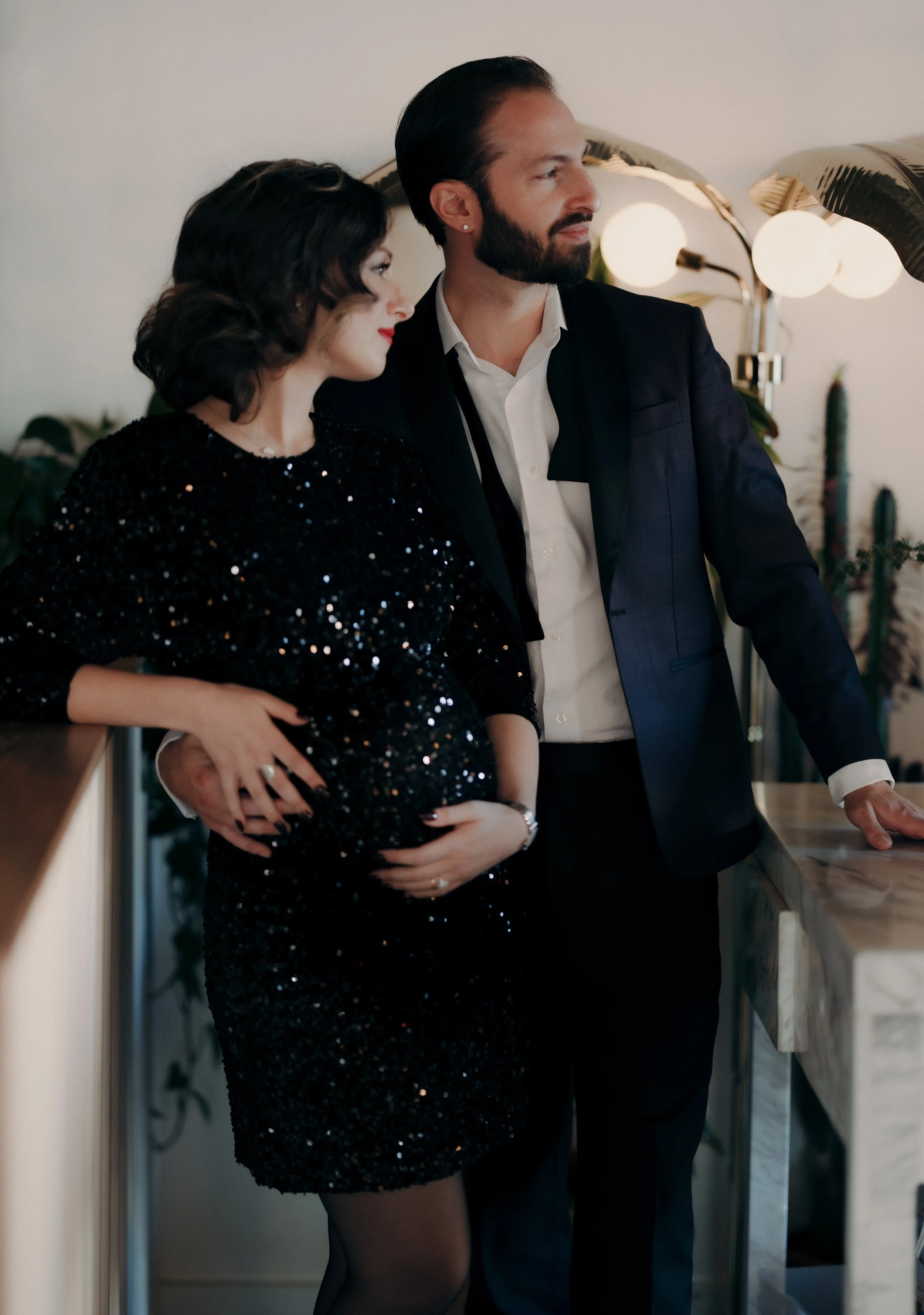 A pregnant woman in a black sequin dress stands beside a man in a dark suit, both indoors in a stylish, modern space with plants and lamp lighting in the background.