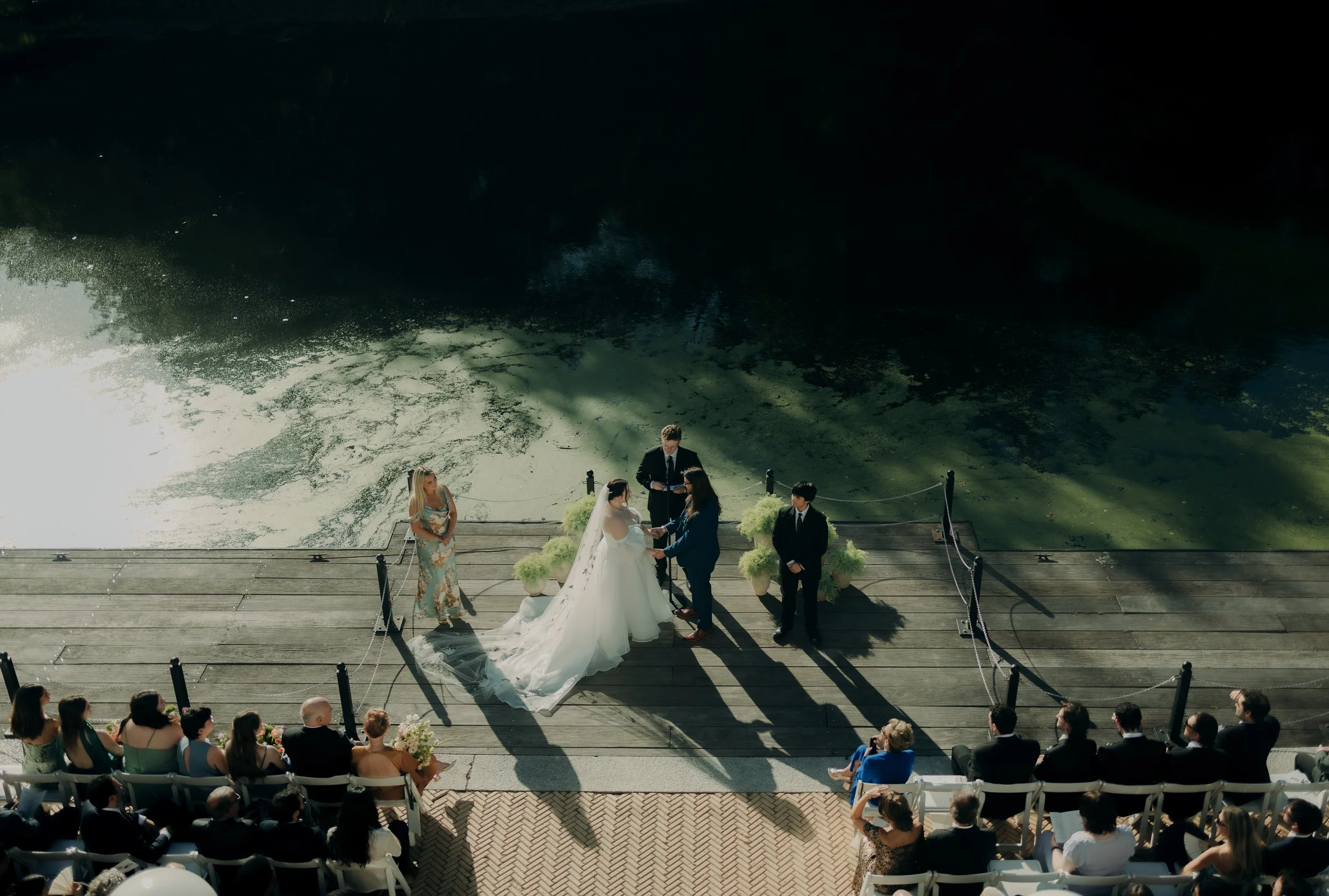 A wedding ceremony taking place on a wooden dock beside a river. The bride and groom hold hands facing each other, with an officiant standing behind a small table. Bridesmaids and groomsmen stand nearby, and guests are seated on white chairs and benches, watching the ceremony. The shadow of the wedding party extends onto the deck.
