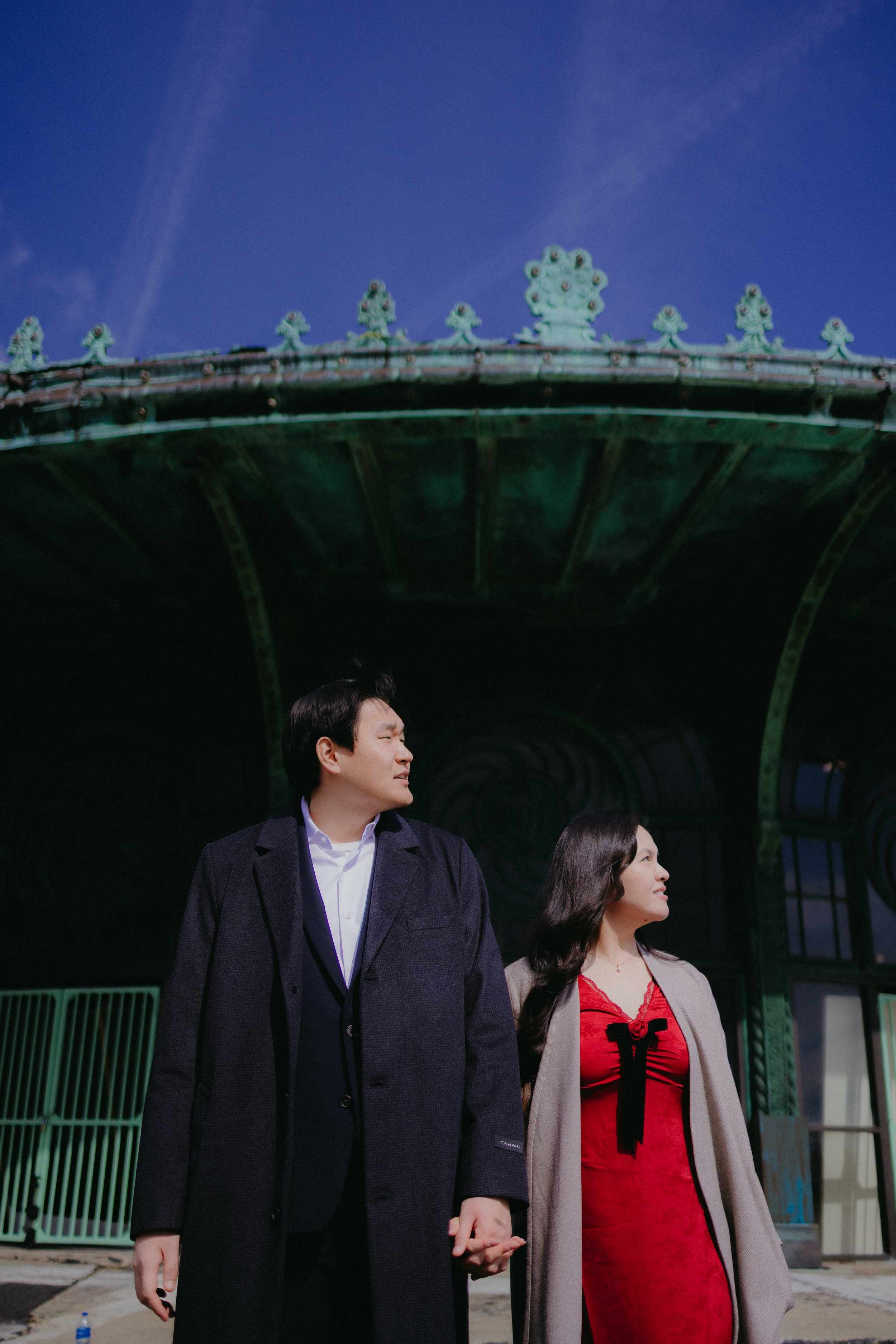 A man and woman holding hands, standing outdoors under a large ornate structure with a blue sky in the background.