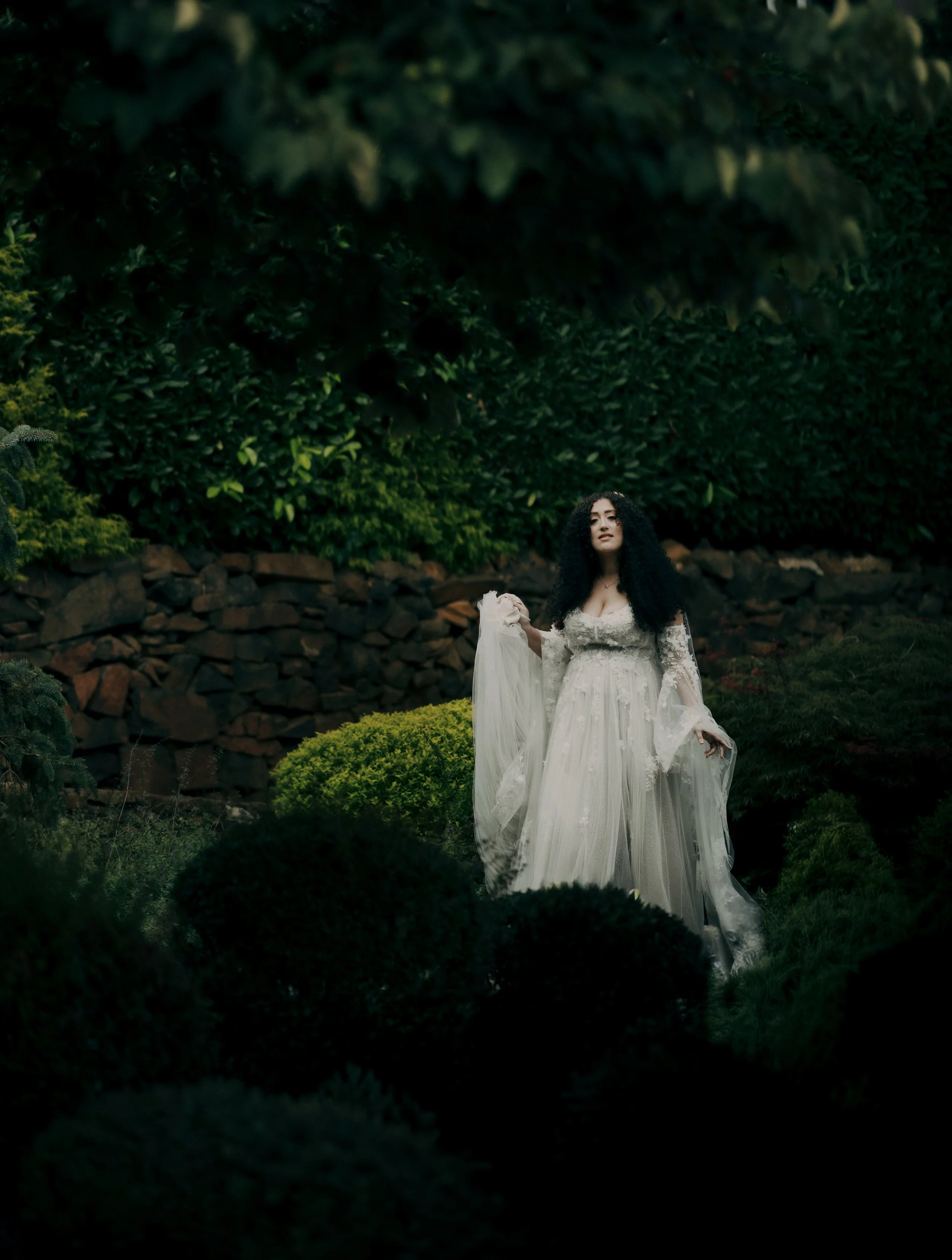 Woman in a flowing white dress standing in a garden with lush greenery and a stone wall in the background.
