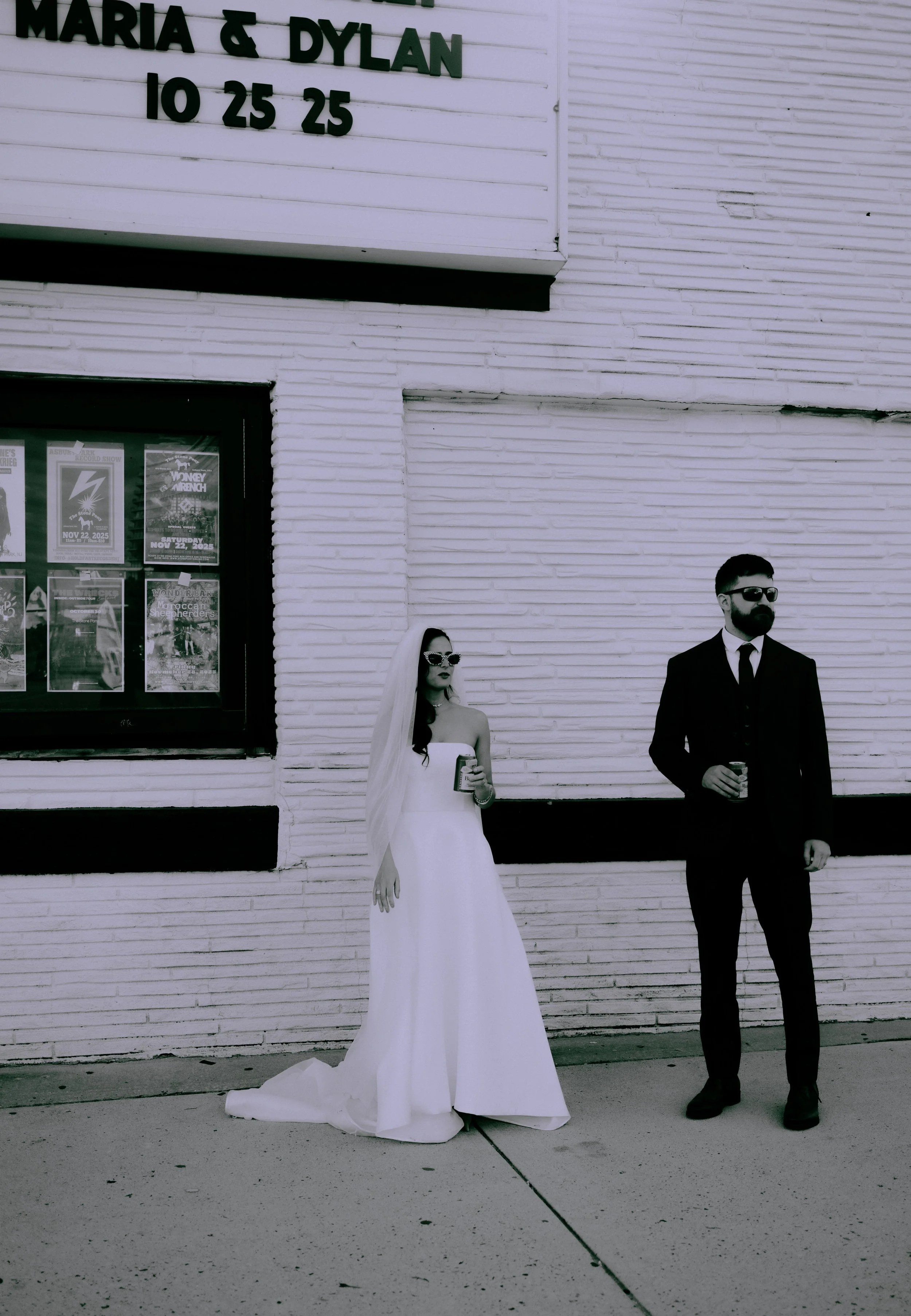 A bride in a white wedding dress and veil stands next to a groom in a black suit and sunglasses outside a white brick building, both holding drinks.