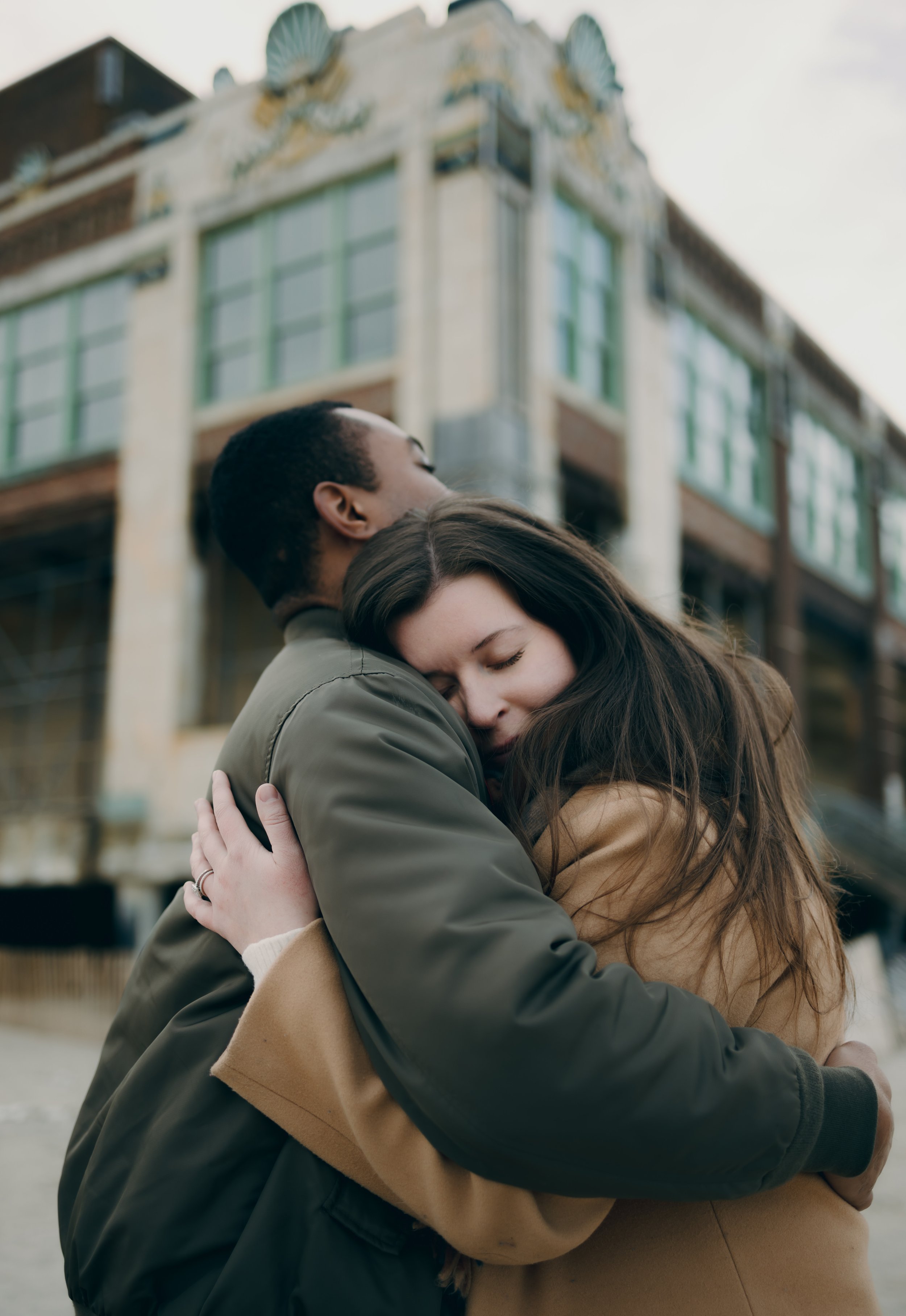 A couple hugging in front of a building with large windows and ornate architecture.