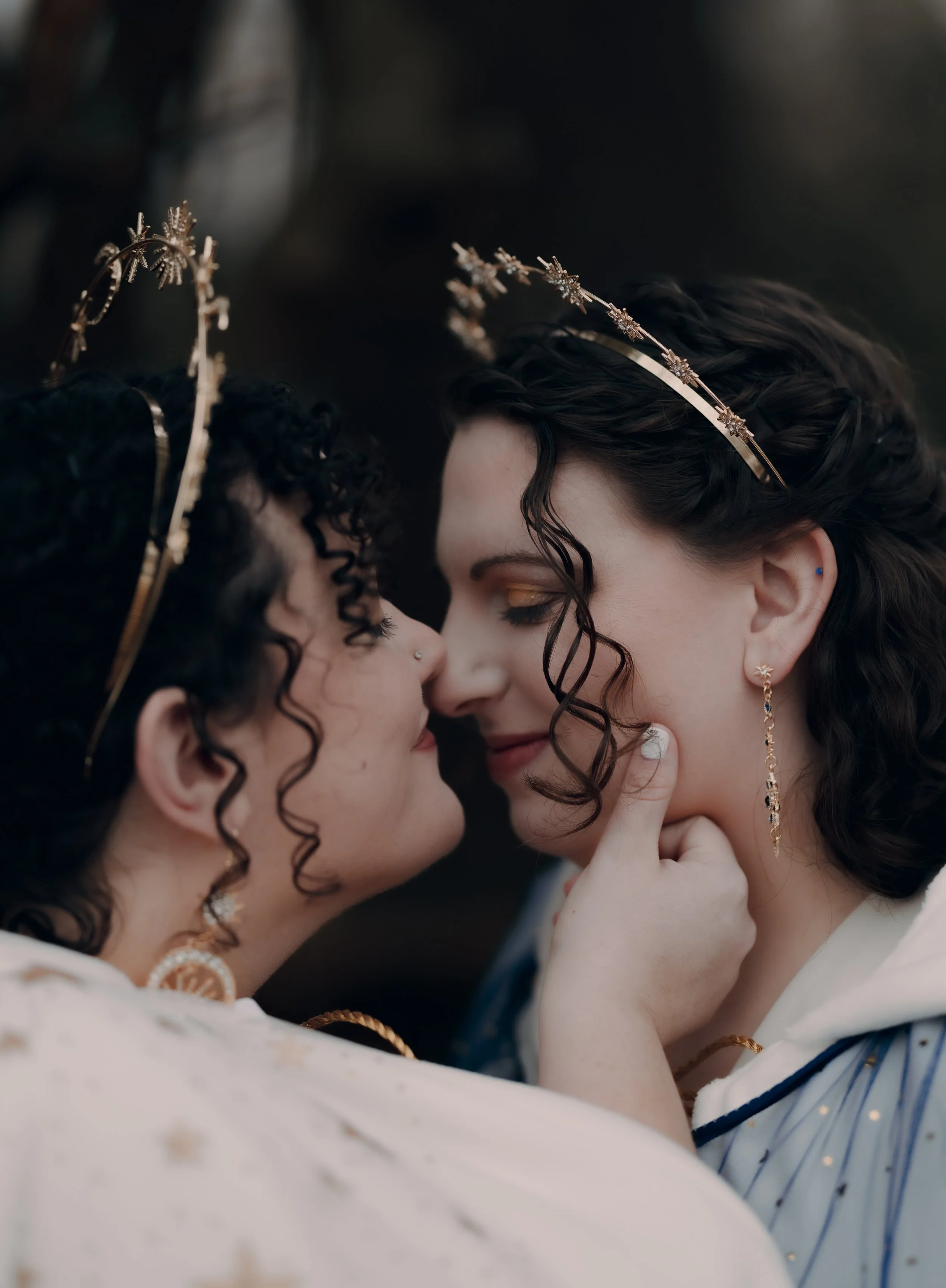 Two women wearing gold tiaras and earrings, touching noses and smiling, with one woman gently touching the other's chin. They have dark, curly hair and are dressed in elegant outfits.