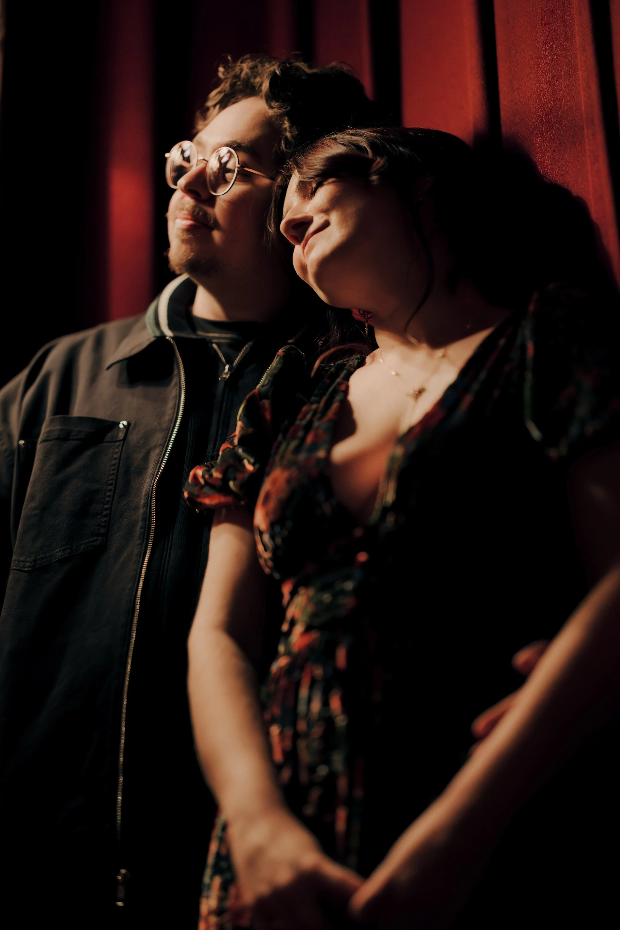 A young man with glasses and a woman with covered eyes stand closely together against a red curtain backdrop, with the woman's head resting on the man's shoulder.