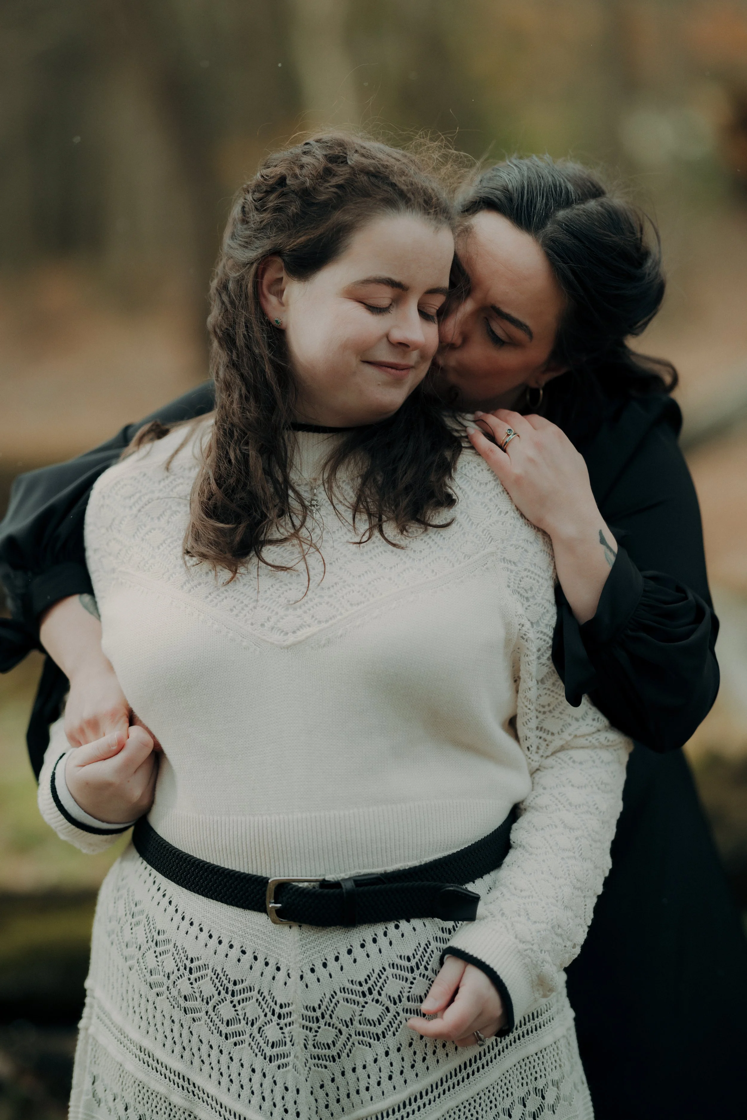 Two women embracing outdoors, one kissing the other's cheek with eyes closed, surrounded by autumn trees.