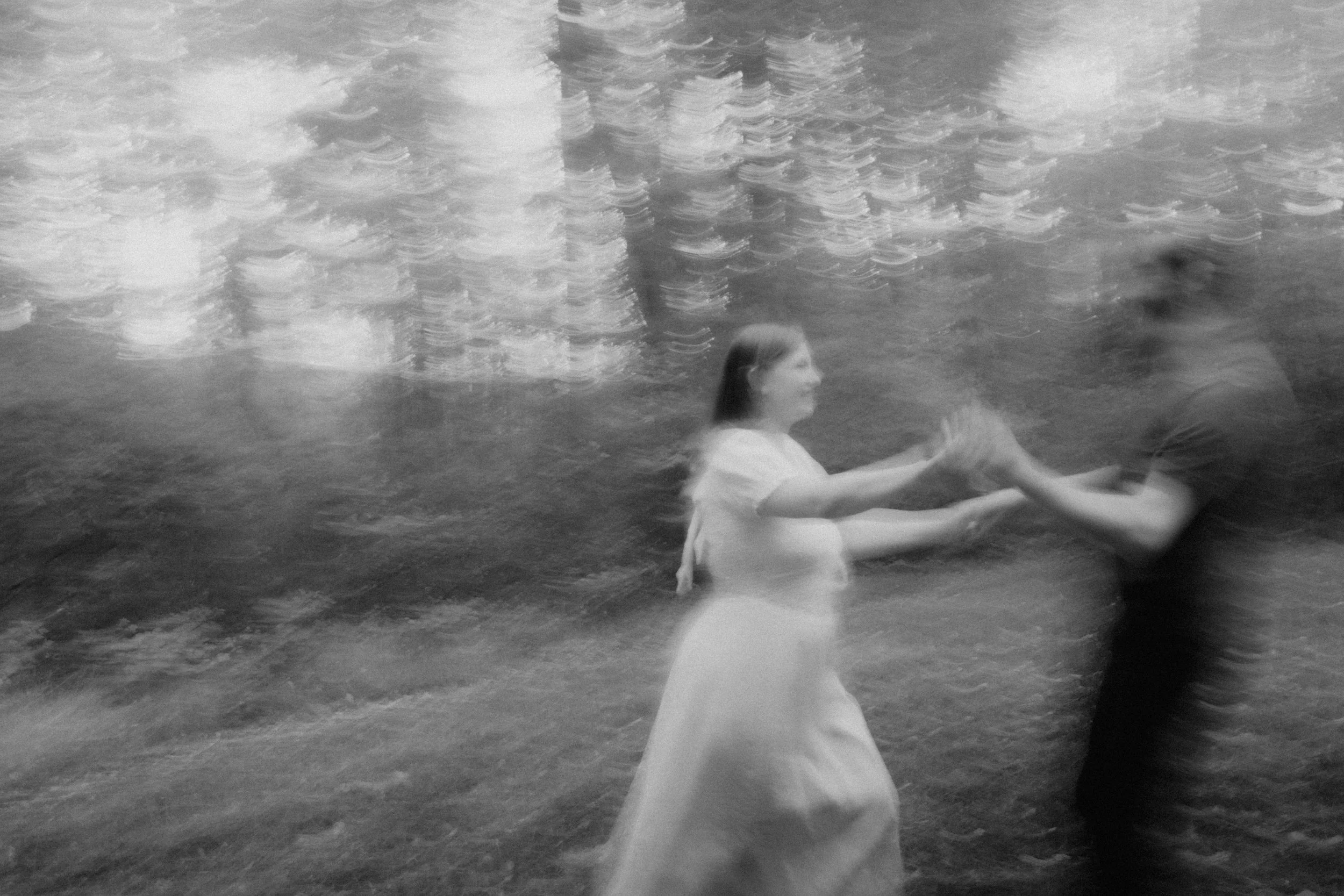 A black-and-white photo of a woman in a light-colored dress dancing with a man in dark clothing in a shallow water body, with a blurred cityscape in the background.