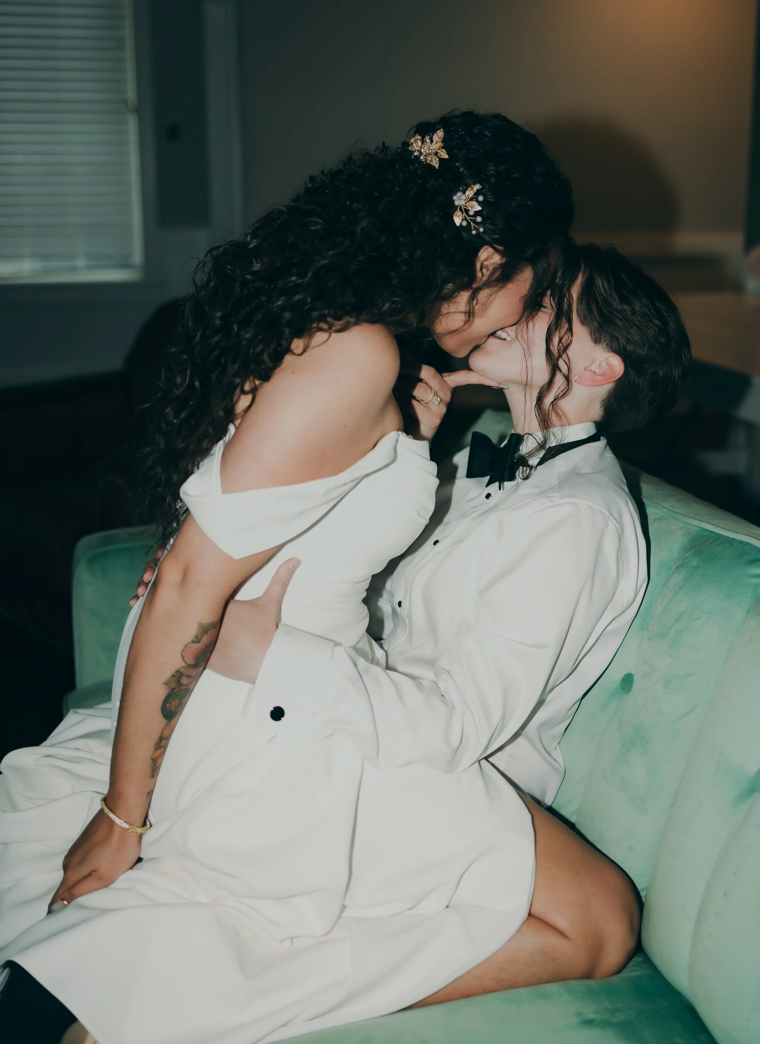 A couple dressed in formal attire sharing a kiss on a green couch in a cozy indoor setting.