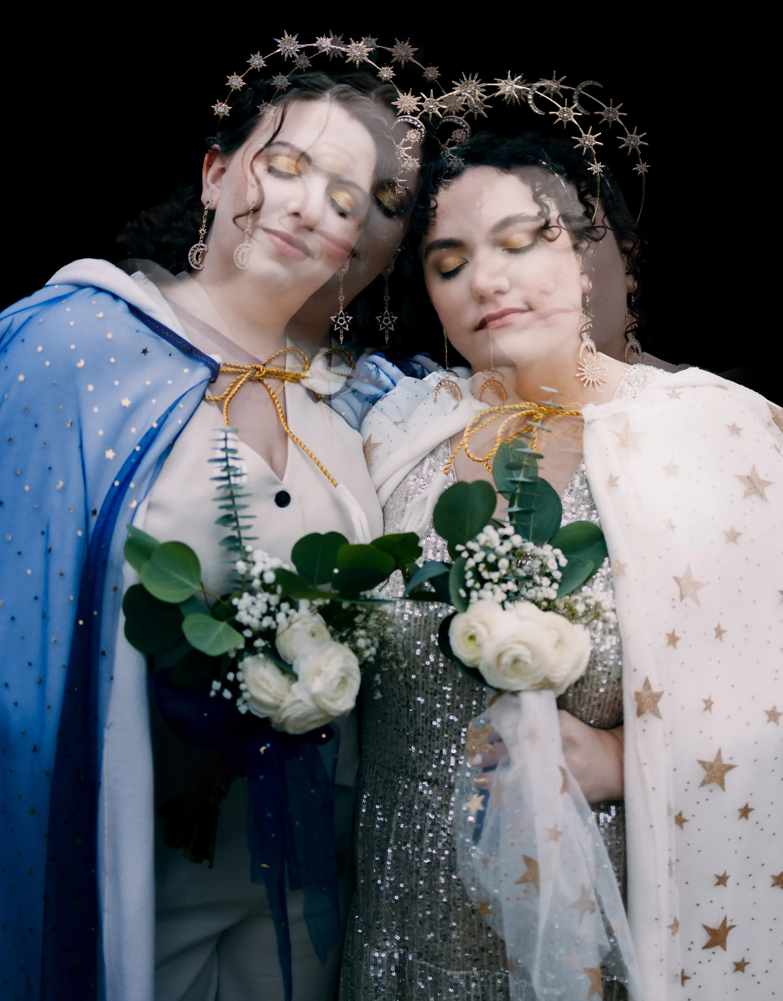 Two women dressed as princesses or fairy tale characters, wearing star and moon themed crowns and capes, holding bouquets of white flowers with greenery,/dressed in sparkly and star-patterned costumes, standing close together with a slight double exp