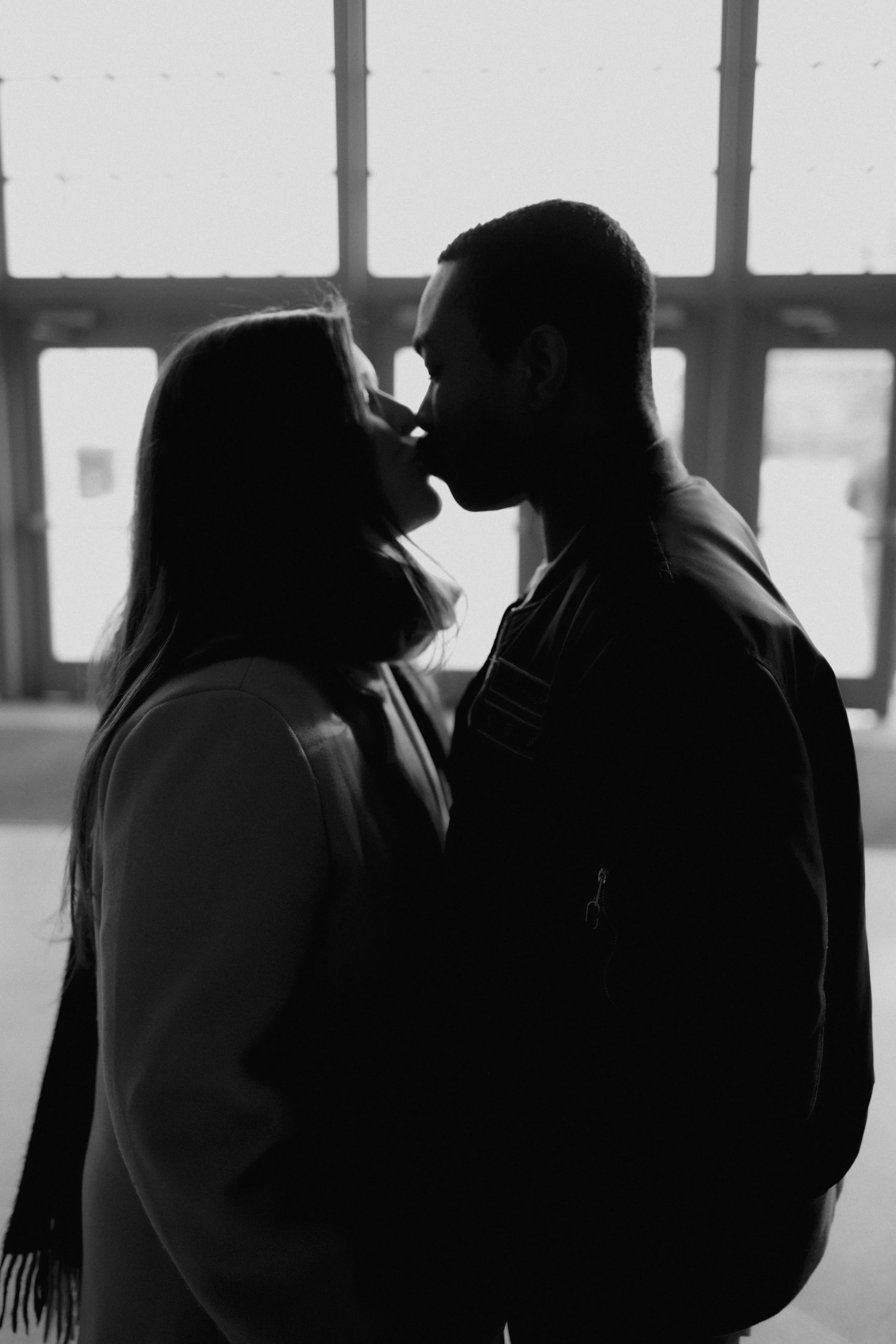 Silhouette of a couple kissing indoors near large windows in black and white.