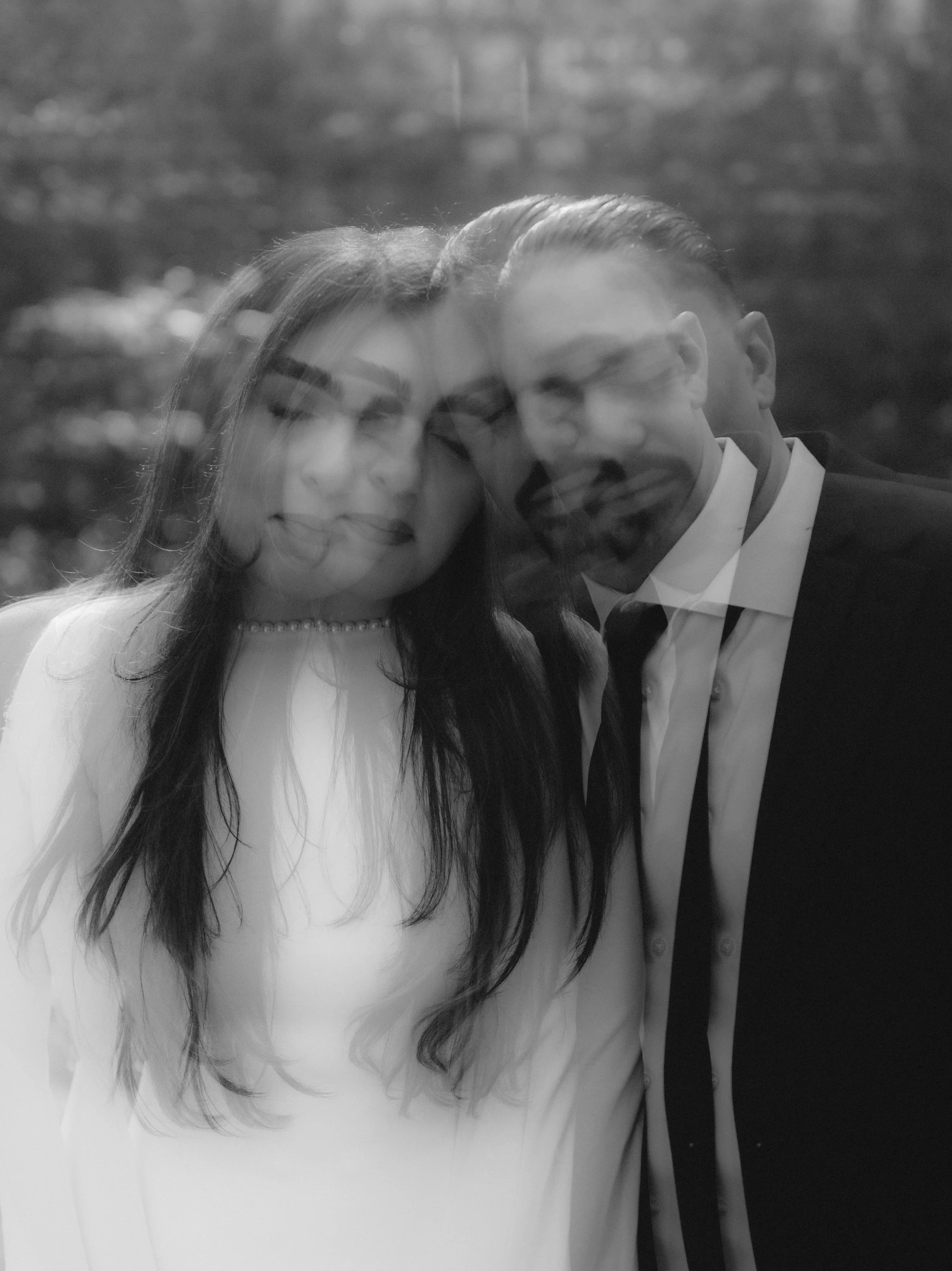 Black and white photo of a couple with their faces overlapping, smiling. The woman has long hair, wears a pearl necklace and a light-colored dress. The man has slicked-back hair, a beard, and wears a suit with a tie. They appear to be outdoors.