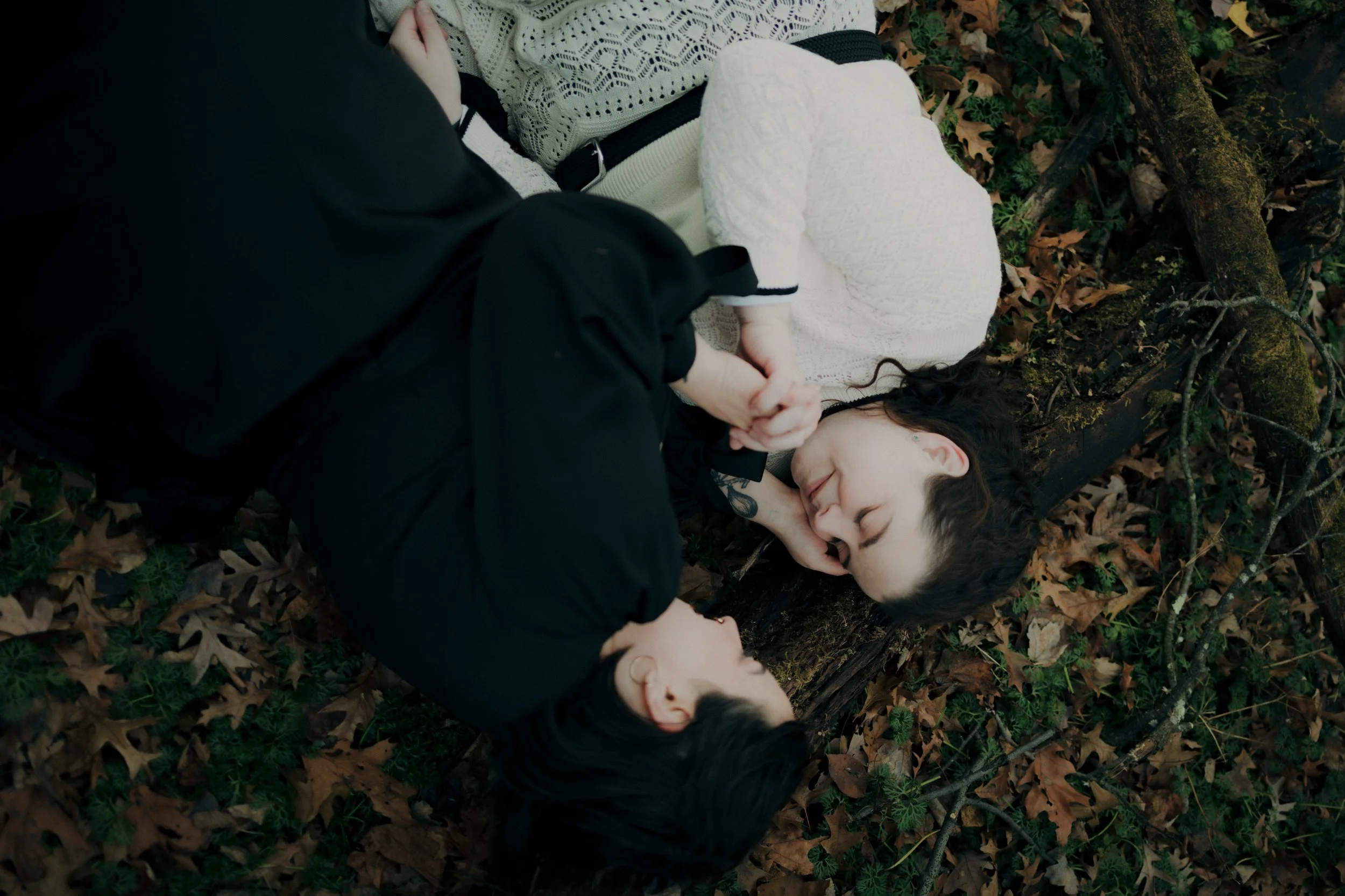 Two women laying on the ground among fallen leaves and branches, holding hands and smiling at each other, outdoors in a forested area.