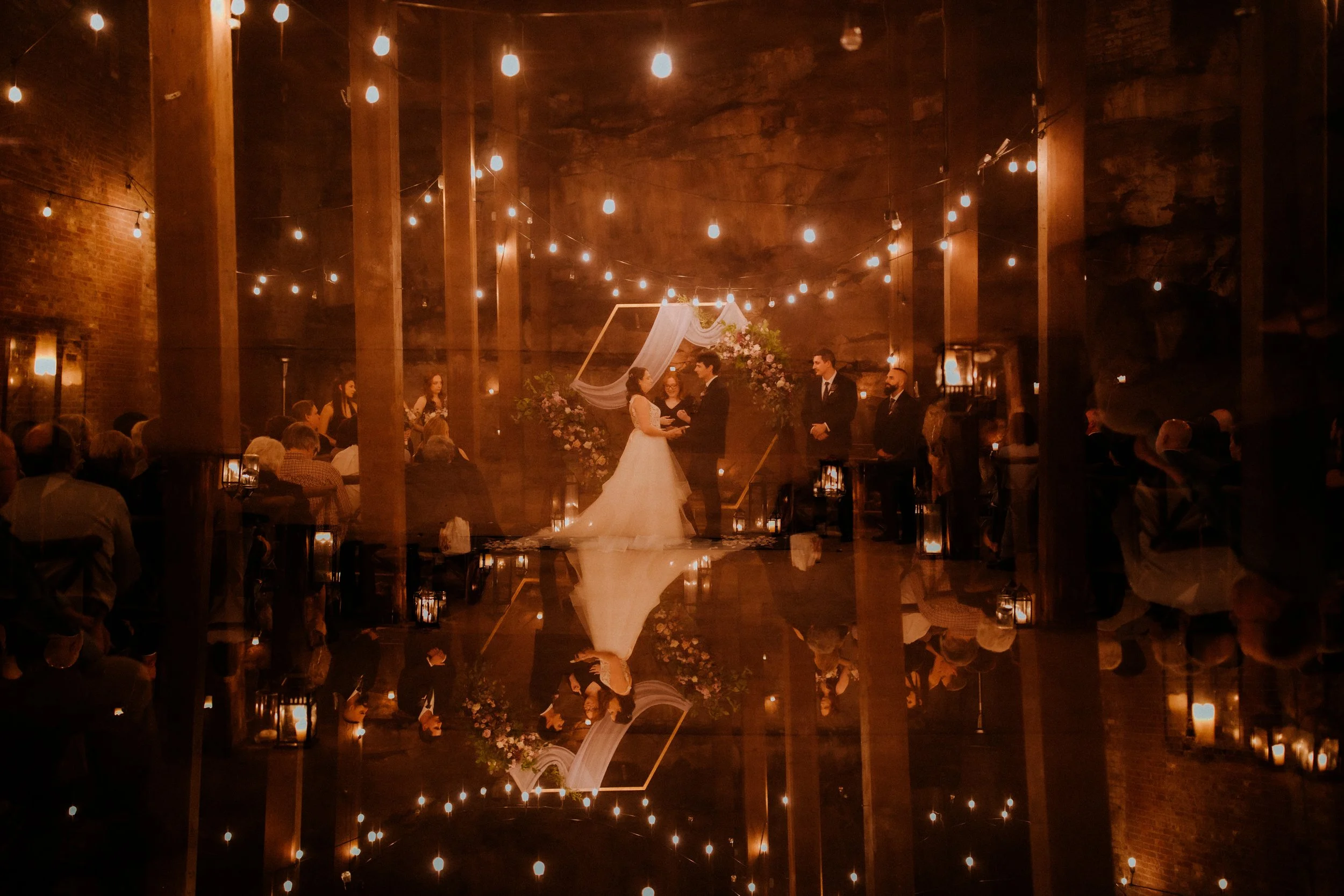 A wedding ceremony taking place in a dimly lit indoor venue with string lights overhead. The bride and groom stand at the altar, holding hands, with guests seated on either side. The reflection of the scene can be seen on a shiny floor or surface.