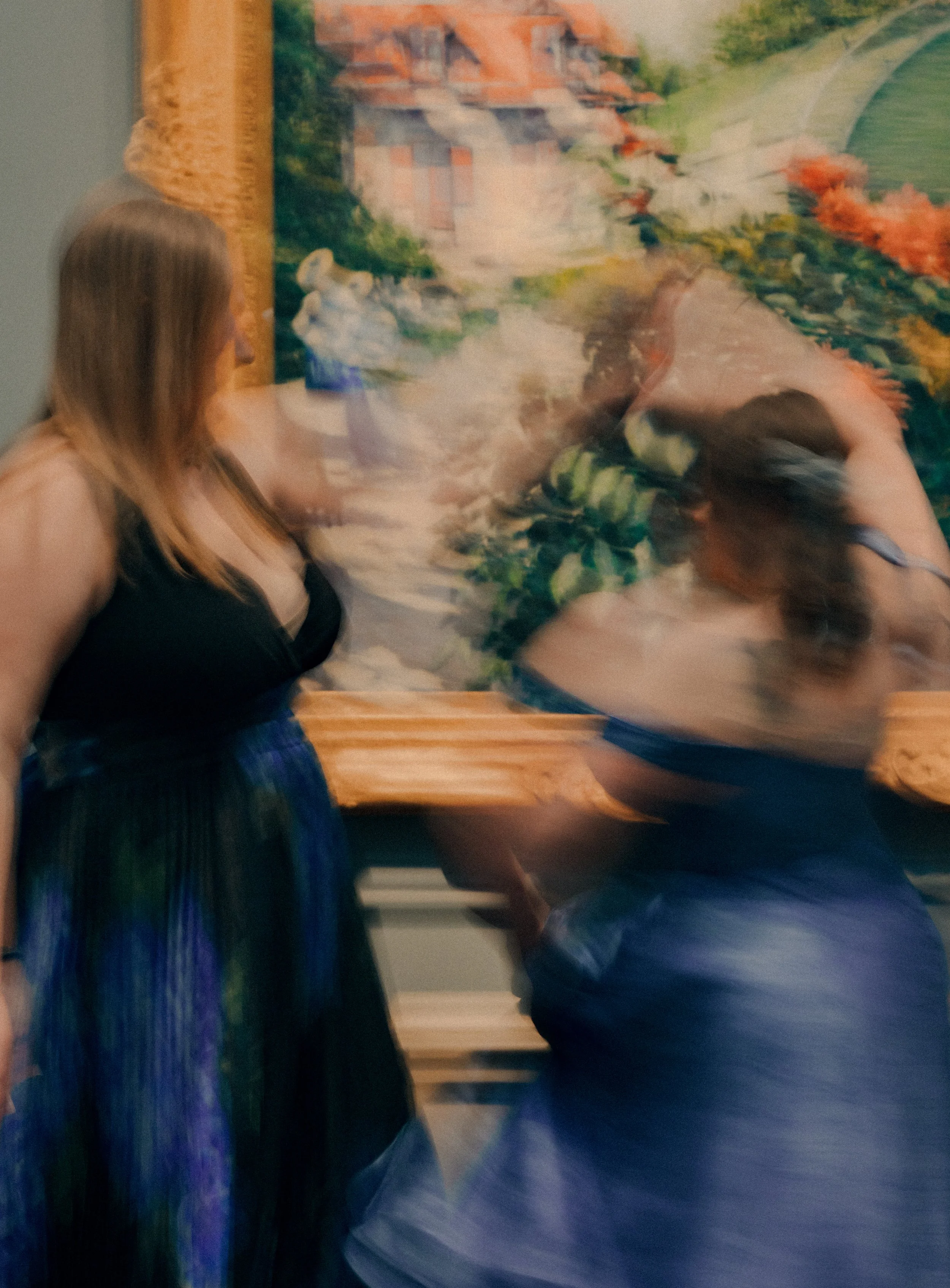 Two women are in front of a colorful painting with a landscape scene. One woman is standing on the left, wearing a black top and a long colorful skirt. The other woman is kneeling or sitting on a bench on the right, wearing a gray top and denim short