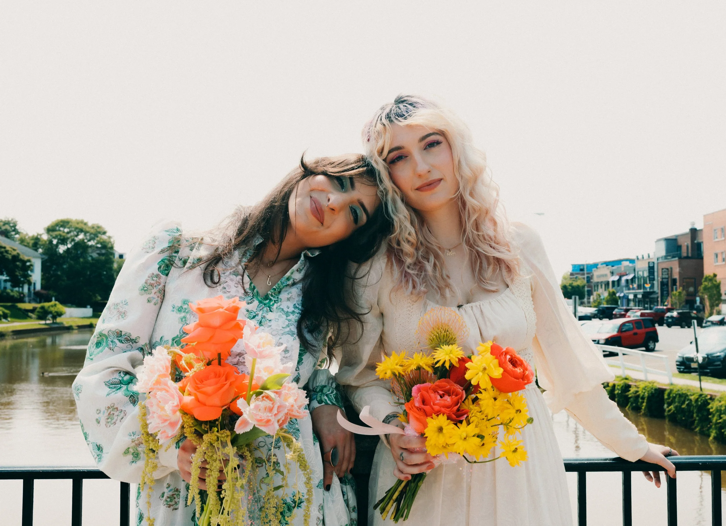 Two women standing outdoors, holding colorful bouquets of flowers, posing near a river with a town in the background on a sunny day.