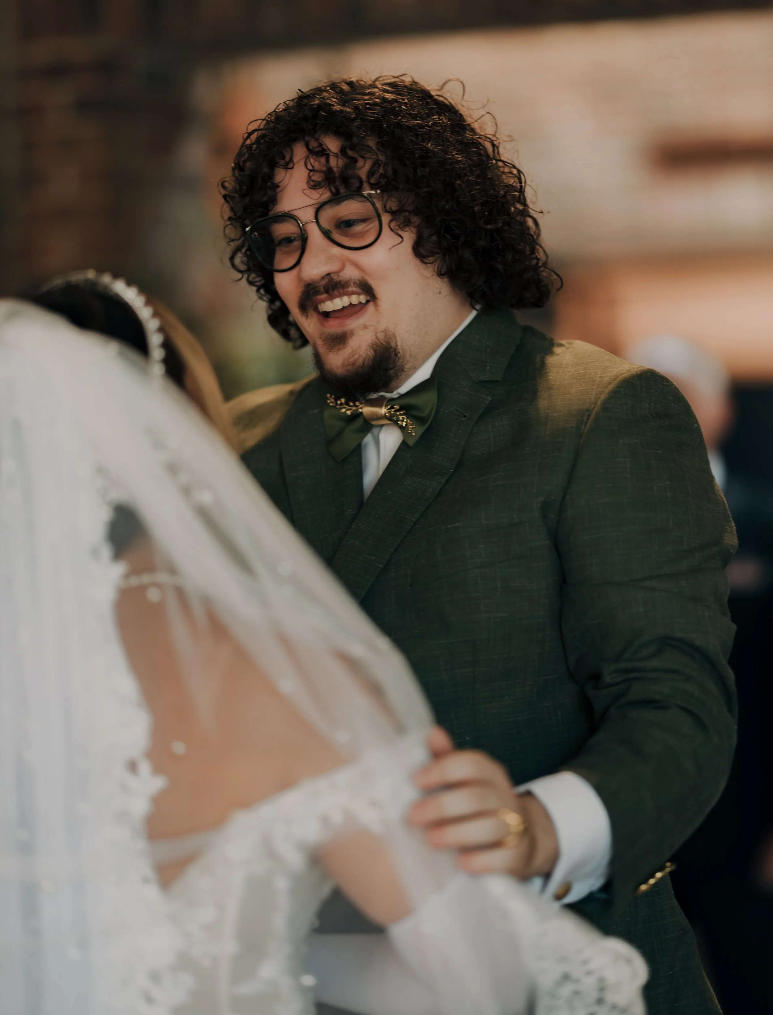 A man with dark, curly hair, glasses, and a goatee, wearing a green suit with a bow tie, smiling and looking at a woman in a wedding dress.