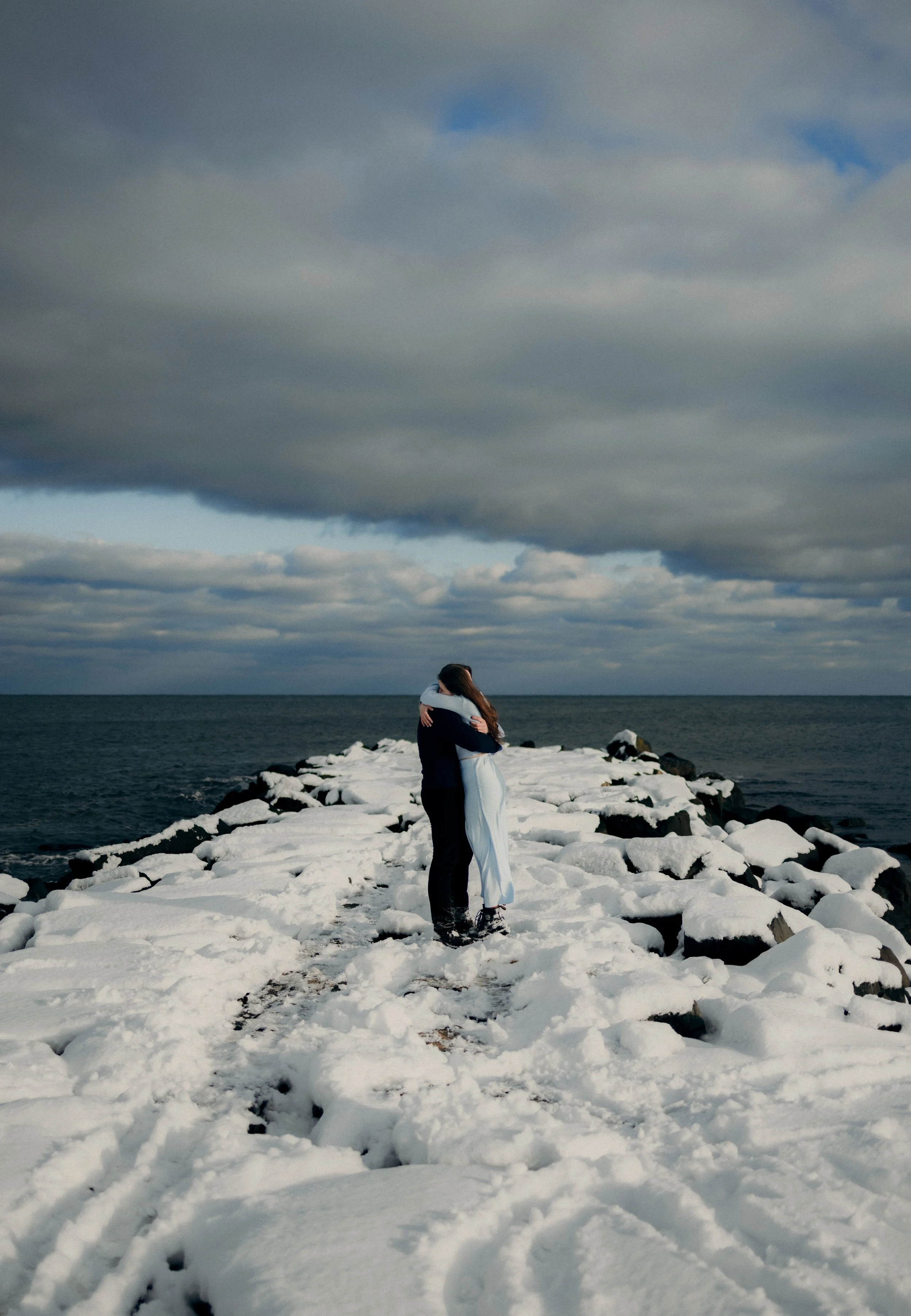 A couple hugging on a snow-covered rocky pier extending into the ocean, with a cloudy sky overhead.