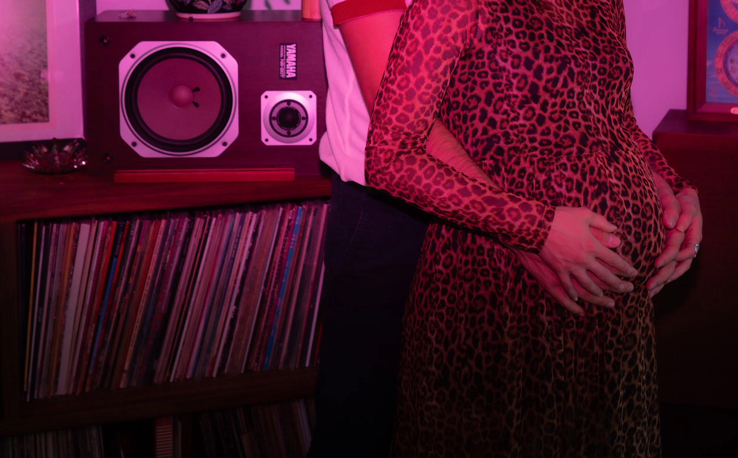 A person in a leopard print dress holding their pregnant belly, with another person standing behind them with a hand on the belly. In the background, there is a shelf with vinyl records and a large speaker on top.