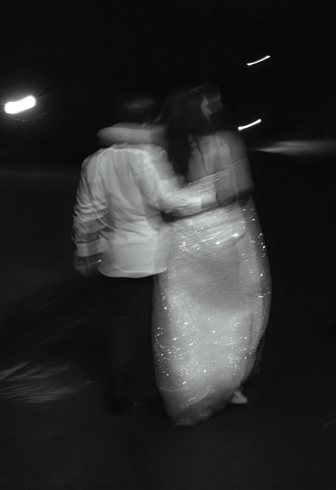 A black-and-white photo of a couple dancing at night, with the woman wearing a glittery dress and the man in a jacket, embracing each other.