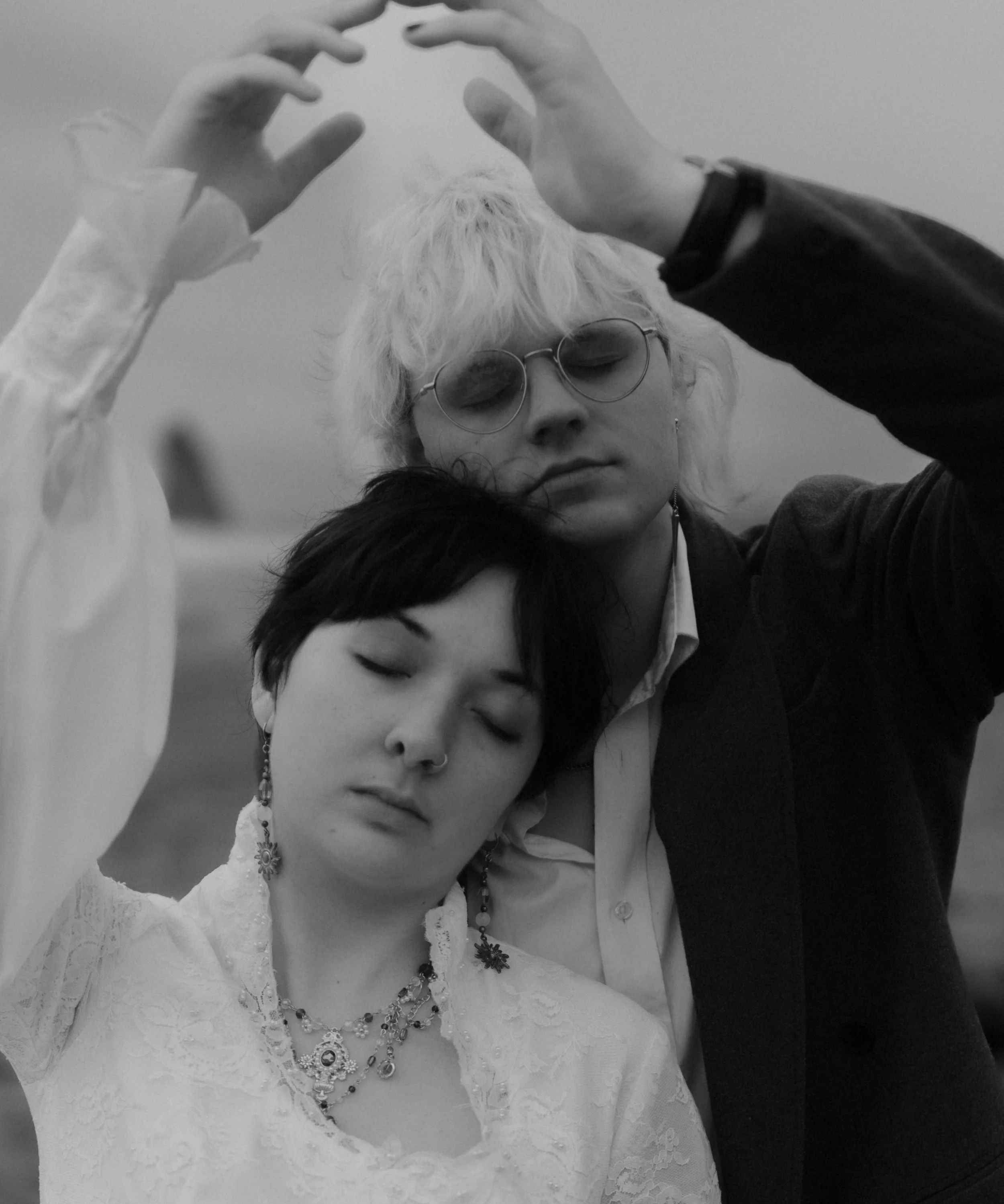 Two women with closed eyes, one with light hair and the other with dark hair, close together with their eyes closed, the light-haired woman making a circular gesture above their heads.