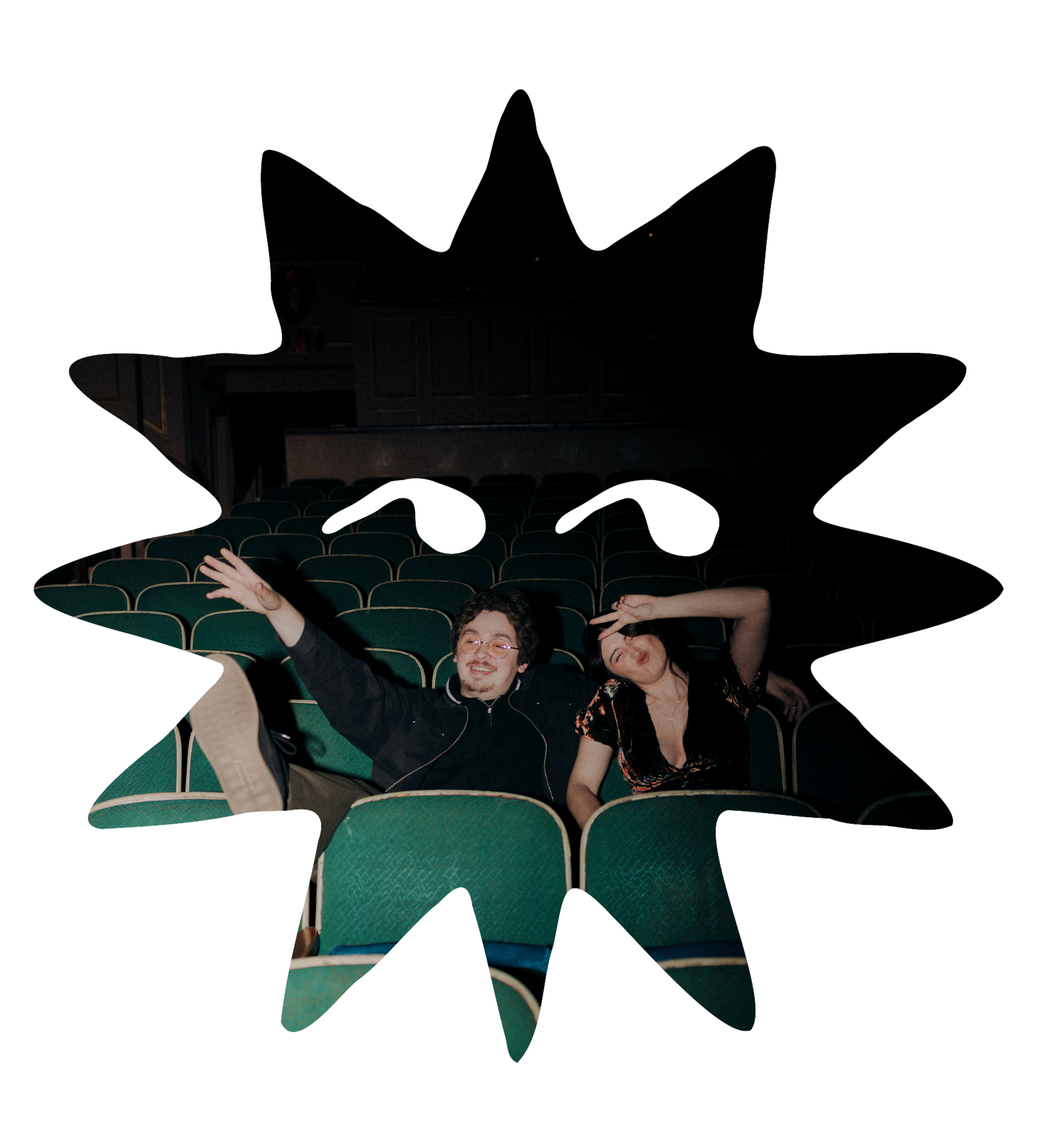 Two people sitting in a nearly empty movie theater, one person smiling and pointing, the other making a peace sign and posing.