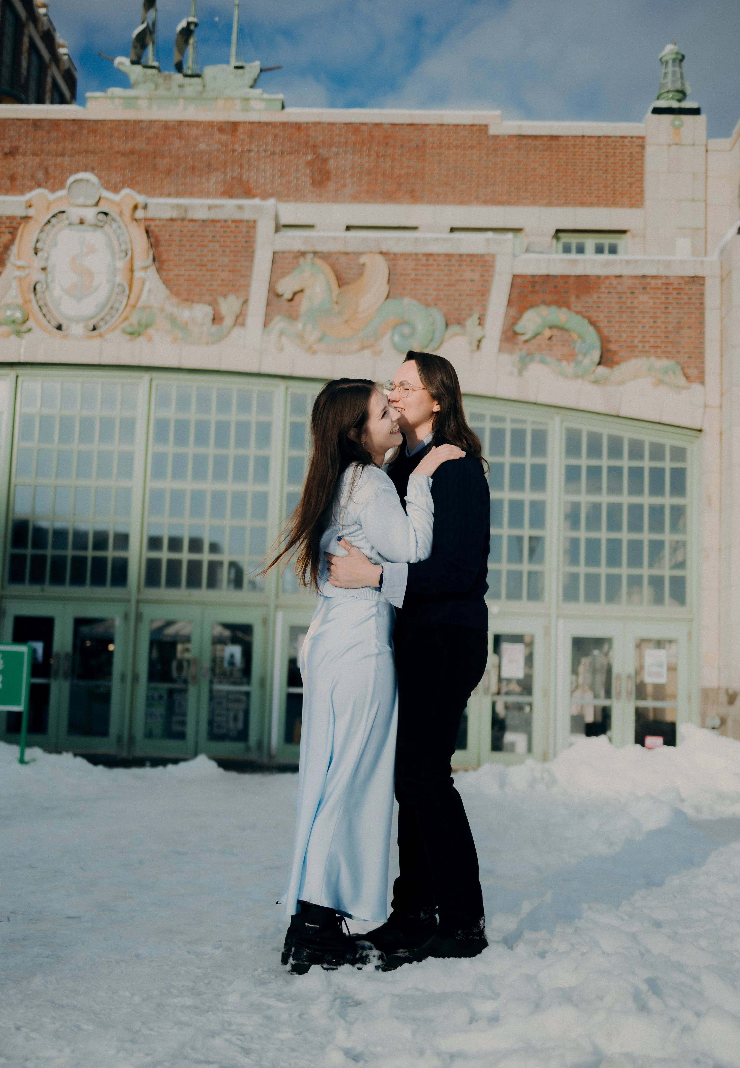 Two women hugging happily in snow in front of a decorative building with large glass windows, a brick wall, and ornate carvings, during daytime.