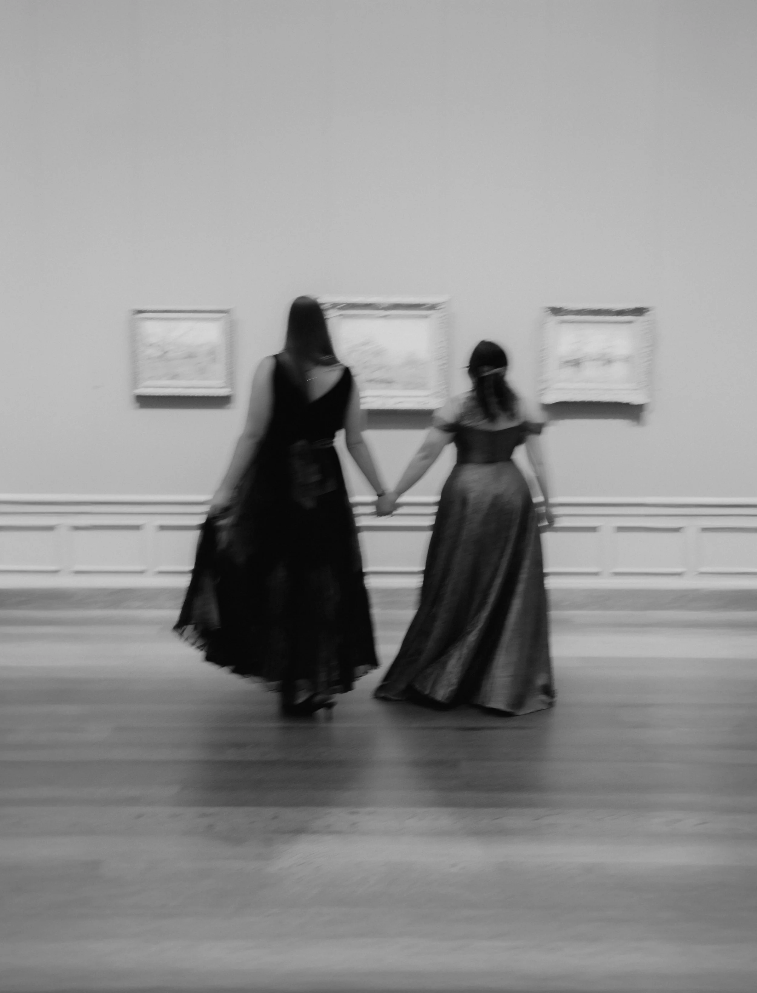 Two women holding hands, walking in an art gallery, wearing long dresses, with framed pictures on the wall behind them.