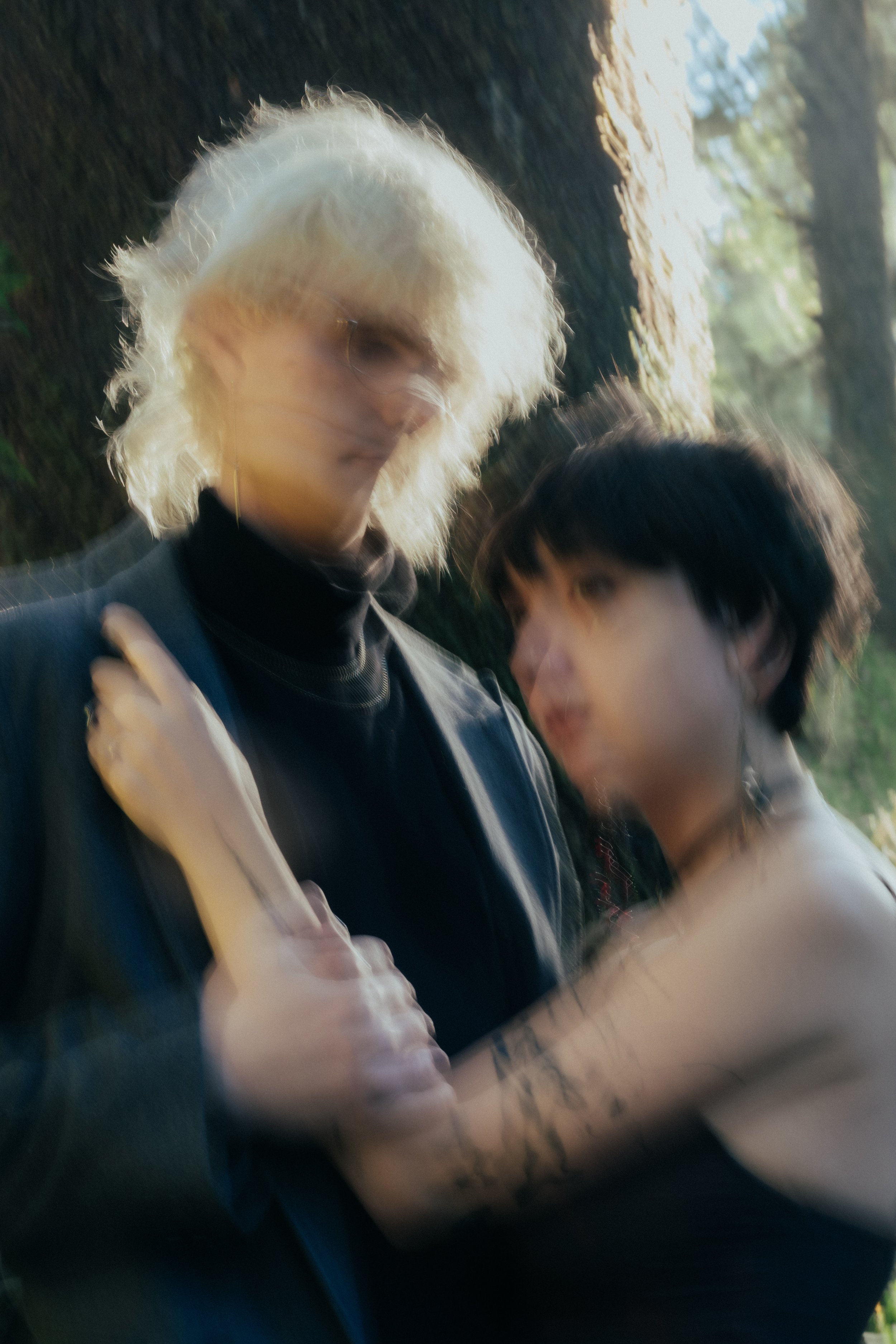 A blurred image of two people, one with platinum blonde hair and the other with dark short hair, standing close together in a natural outdoor setting.
