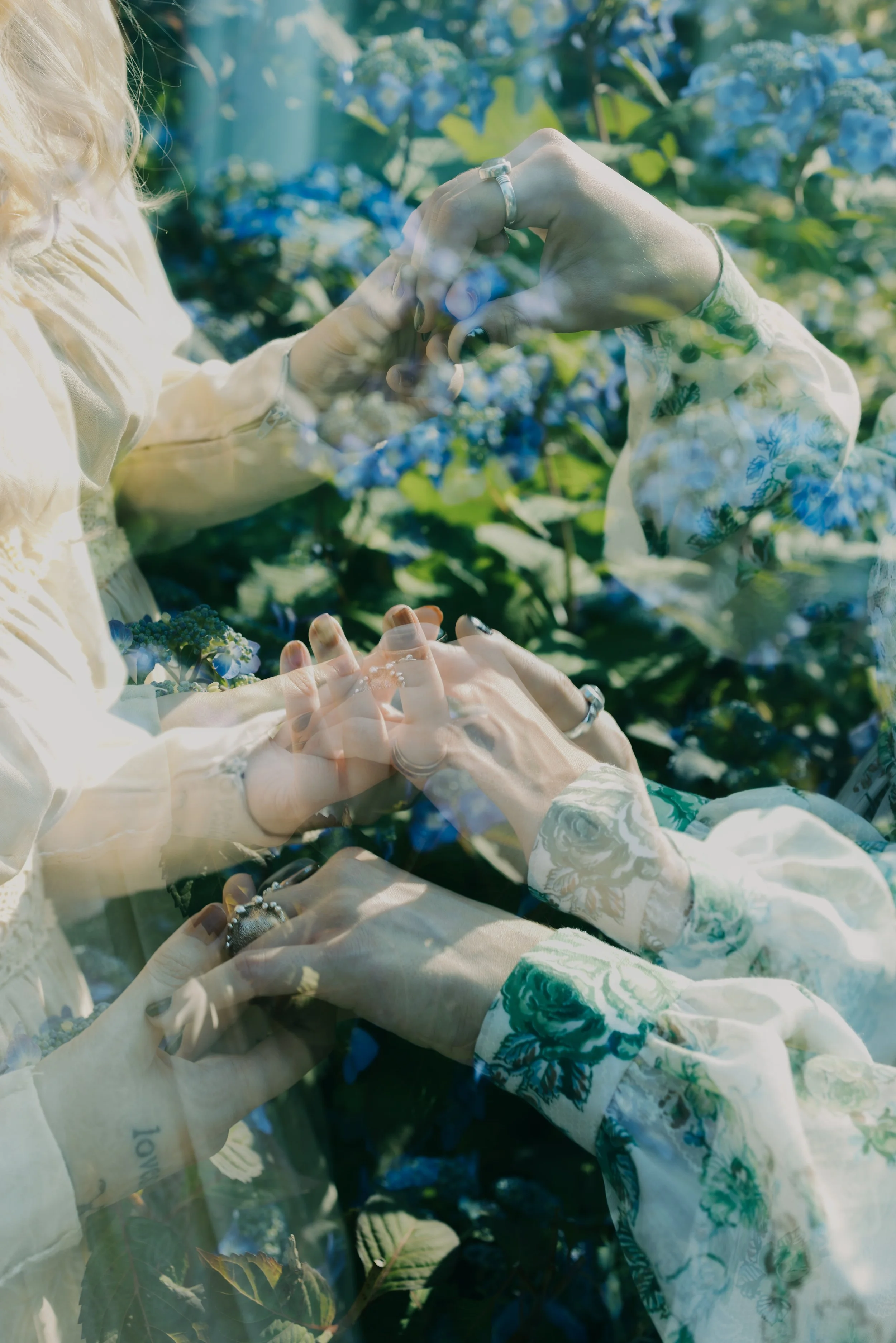 Double exposure photo of two women holding hands among blue hydrangea flowers, with one woman's face partially visible, wearing floral clothing and rings, in a bright outdoor setting.