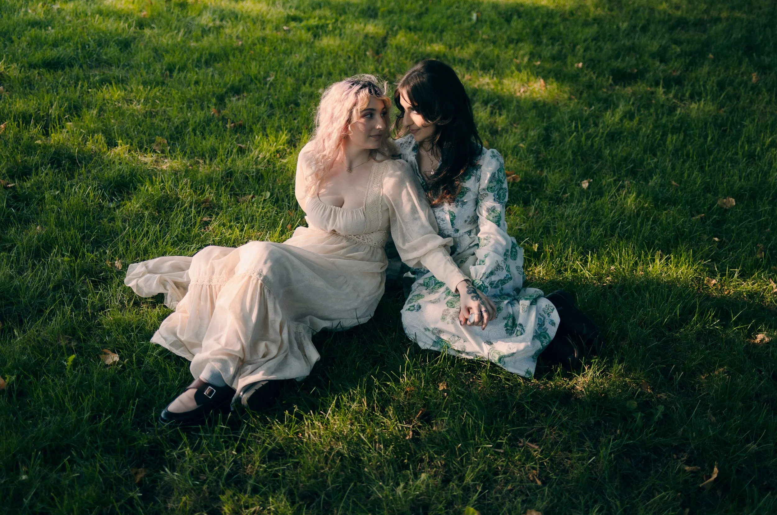 Two women sitting on the grass, facing each other and gazing into each other's eyes, in a park during daytime.