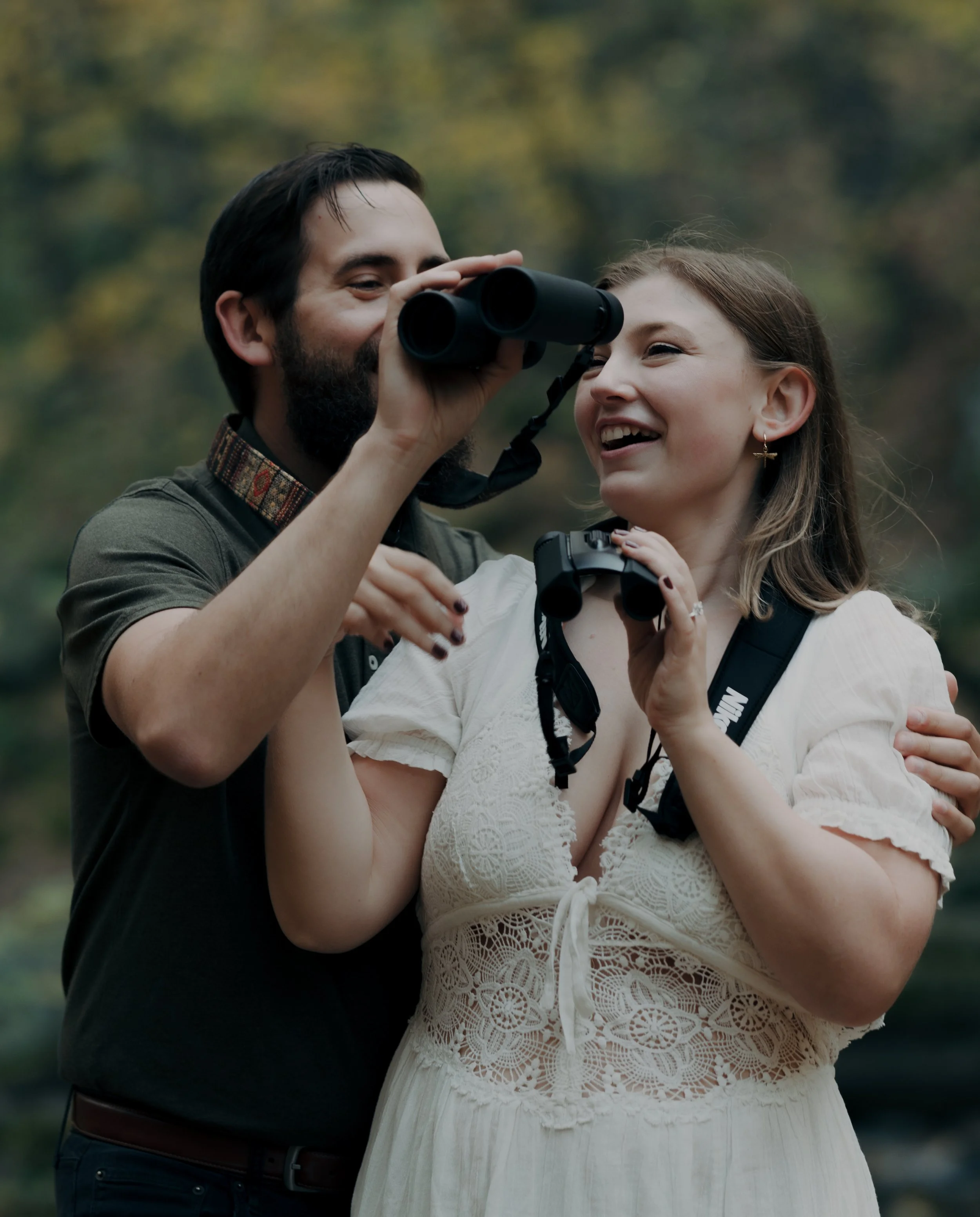 A bearded man and a woman with a camera around her neck stand outdoors, smiling as they look through binoculars, its leaves blurred in the background.