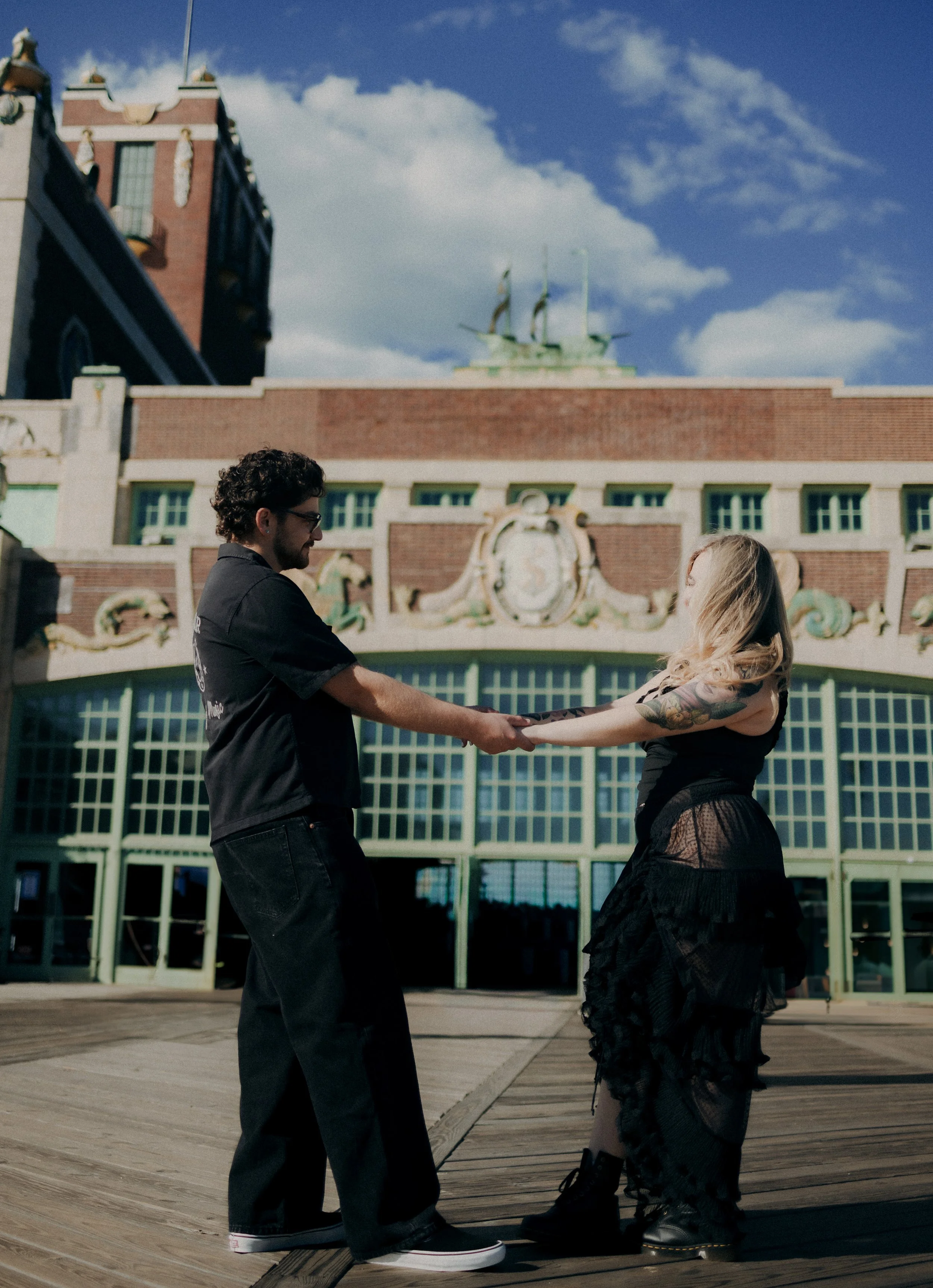 A couple holding hands and dancing outdoors in front of a historic building with decorative details and large windows, under a partly cloudy sky.