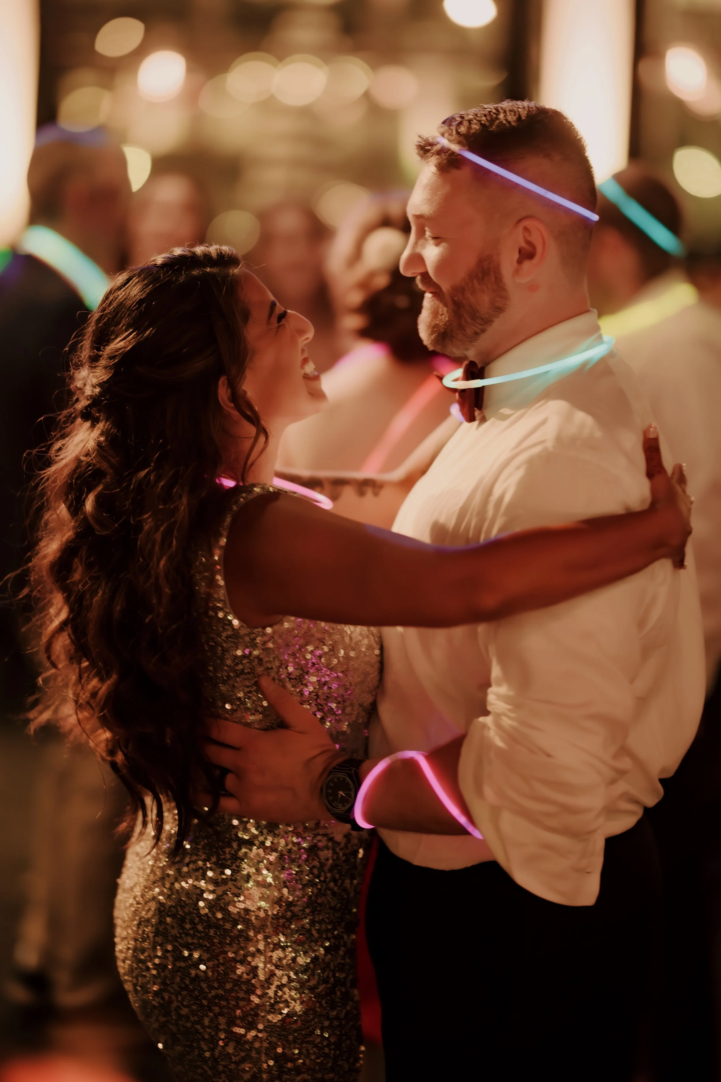 A couple dancing at a party or wedding, with colorful glow sticks around their necks, smiling and looking at each other in a lively, decorated social setting.