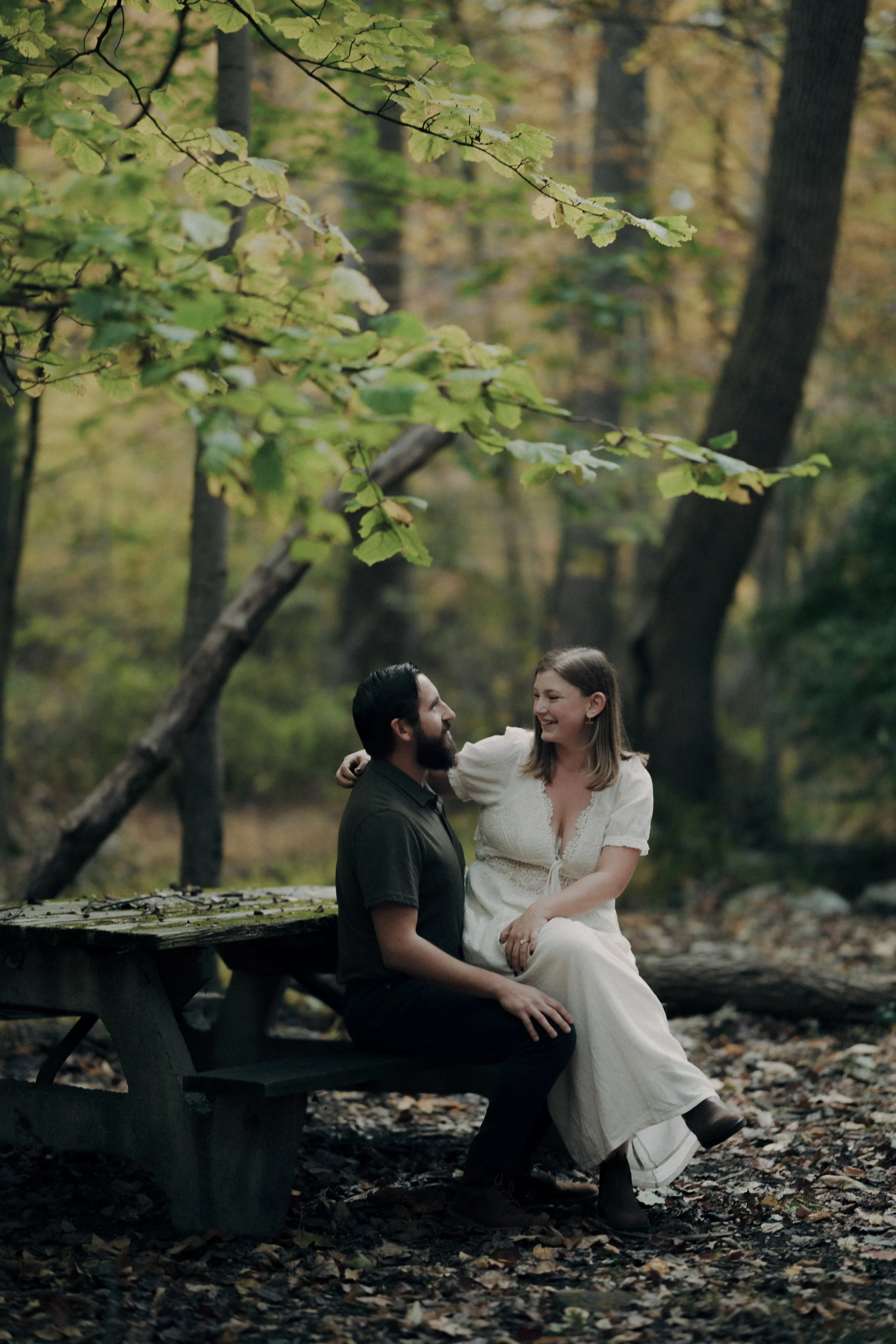 A man and woman sitting on a park bench in a forest, smiling at each other, with fallen leaves around them and trees with green and yellow leaves in the background.